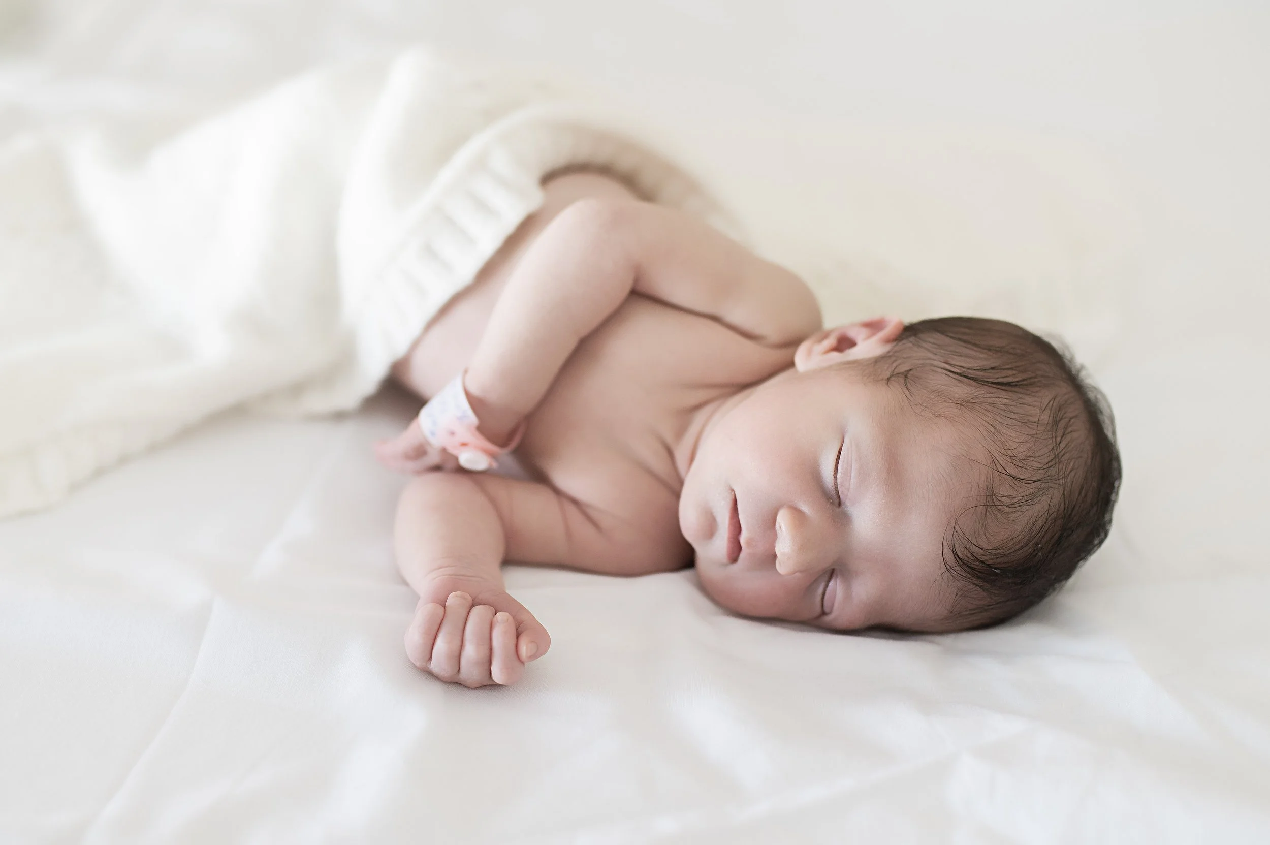 Un bébé endormi sur un lit blanc, partiellement couvert d'une petite couverture blanche.