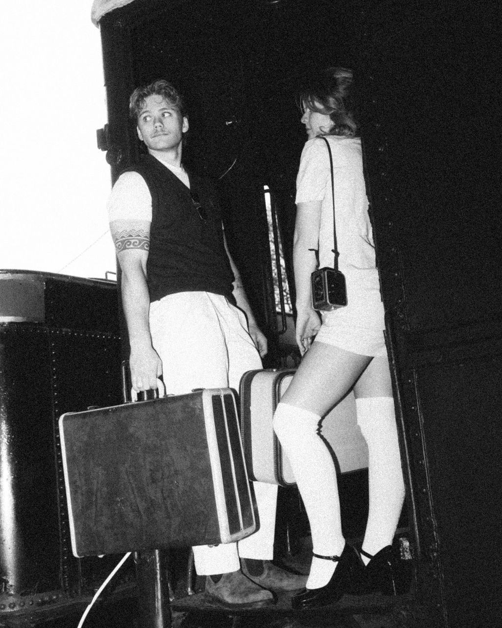 A black and white photo of a young man and woman at an airport or train station. The man is holding a vintage suitcase and looking slightly away, while the woman, wearing thigh-high socks, is holding a smaller suitcase and has a camera hanging around her neck. They are standing near an open doorway or exit.