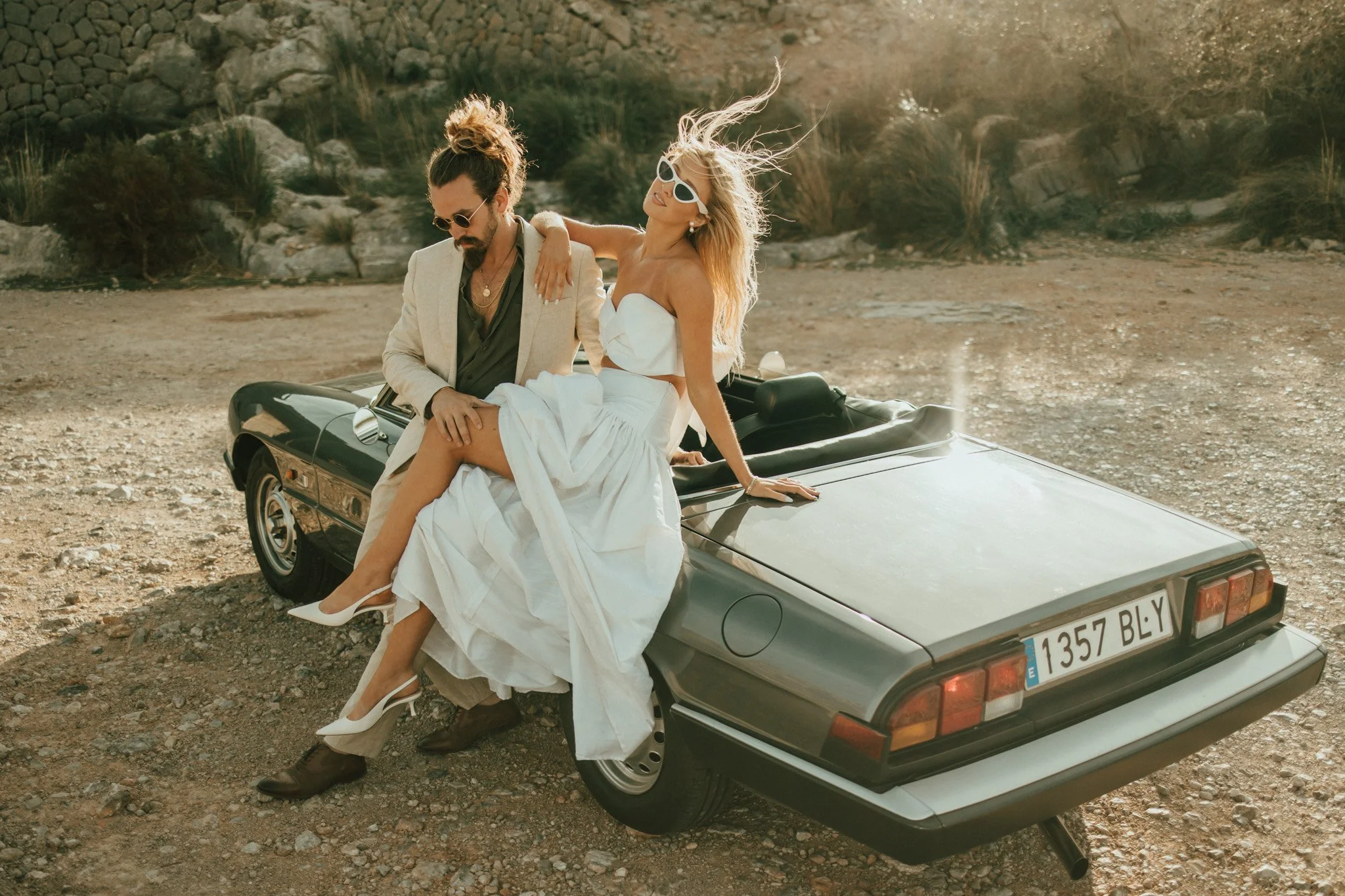 A man and woman sitting on a convertible car in a desert landscape. The woman is wearing a white dress and white heels, while the man is dressed in a beige blazer and dark shirt with boots. The woman is wearing sunglasses and leaning back with her arm on the man, who is looking down.