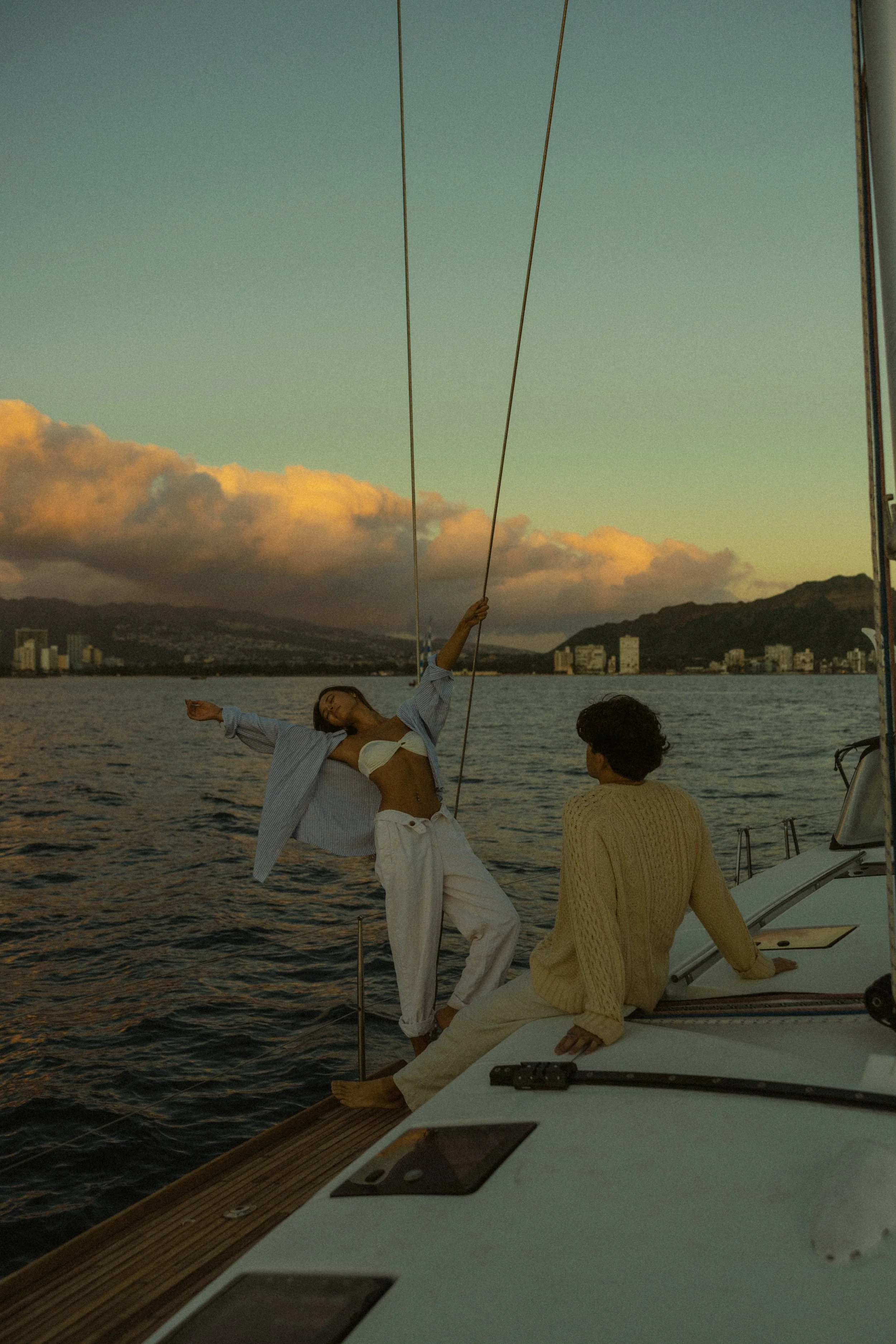 A woman dancing on the deck of a boat at sunset, with a man sitting nearby, overlooking the water and city skyline.