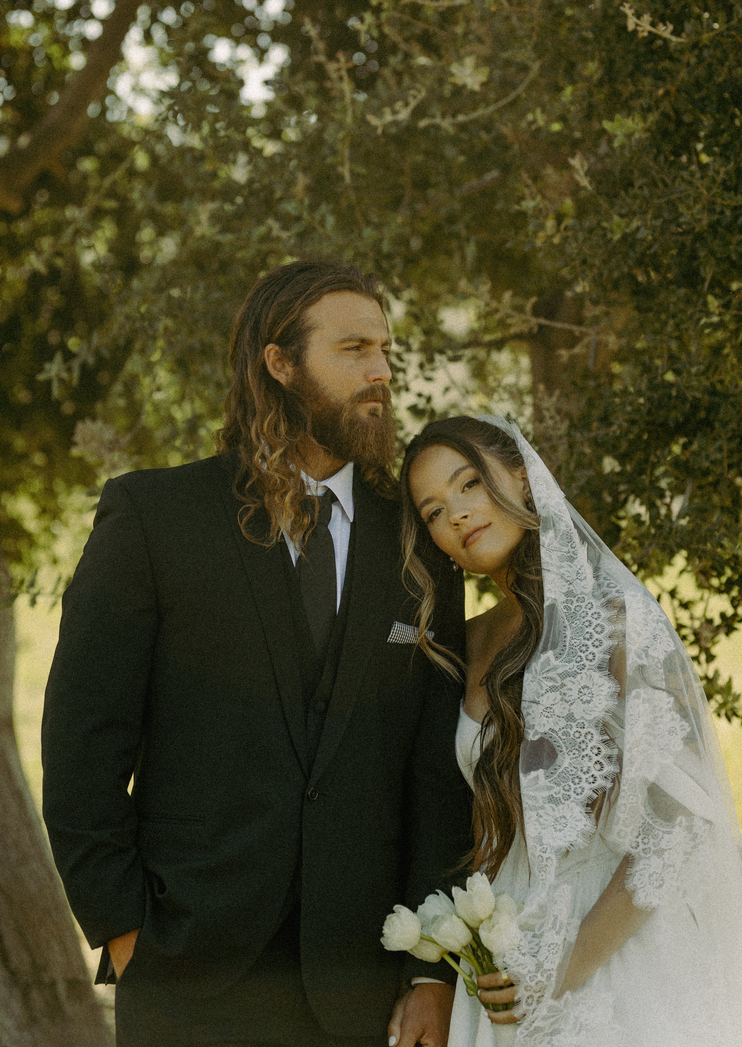 A bride and groom pose outdoors under a tree for a wedding photo. The groom has long hair and a beard, wearing a black suit, and the bride has long dark hair with loose curls, a lace veil, and is holding a bouquet of white tulips.