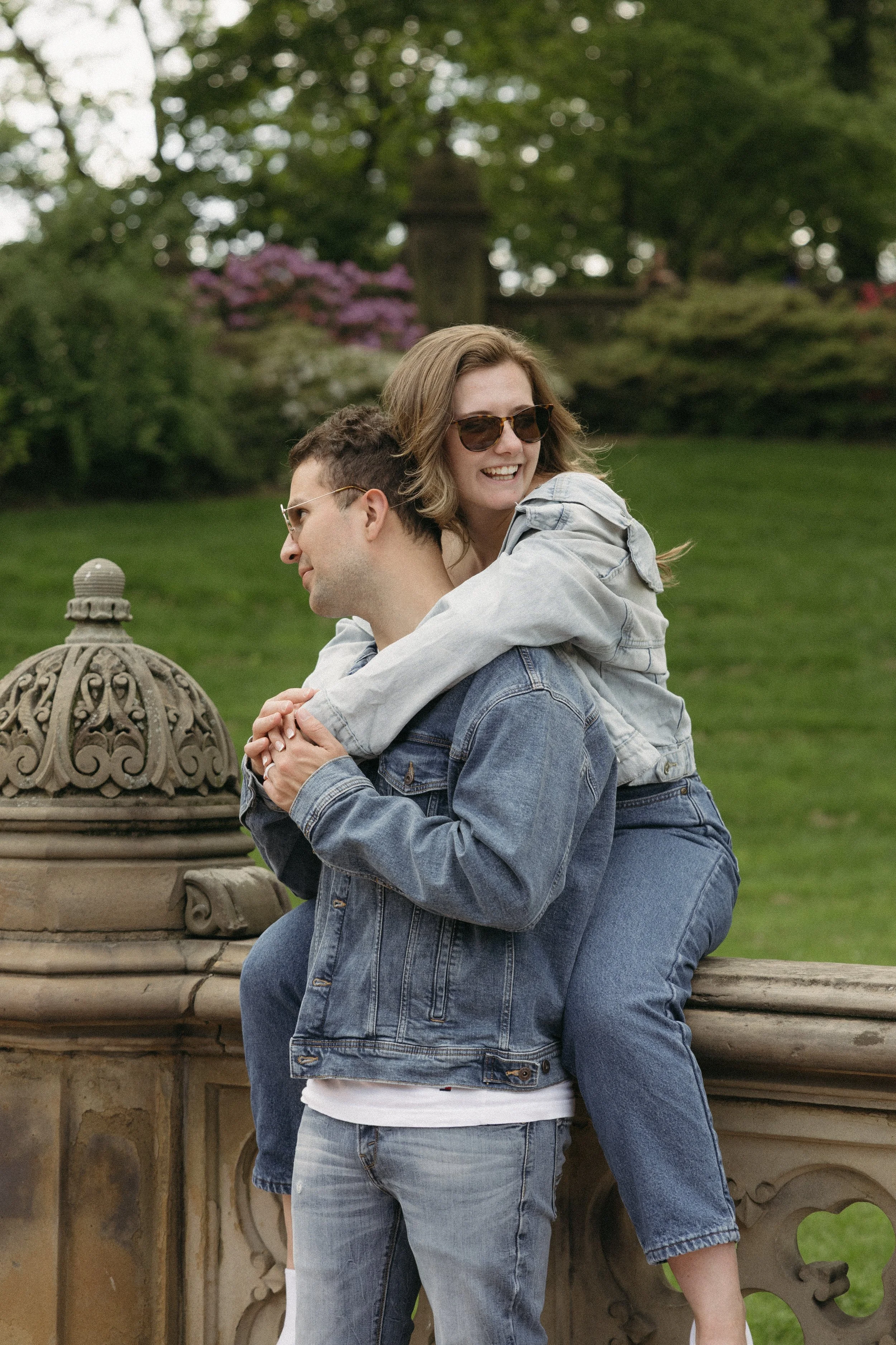 A young couple enjoying time outdoors on a park ledge. The woman, wearing sunglasses, is on the man's back, smiling and embracing him, while the man, in a denim jacket and glasses, stands with his hands clasped. The background features lush greenery and trees.