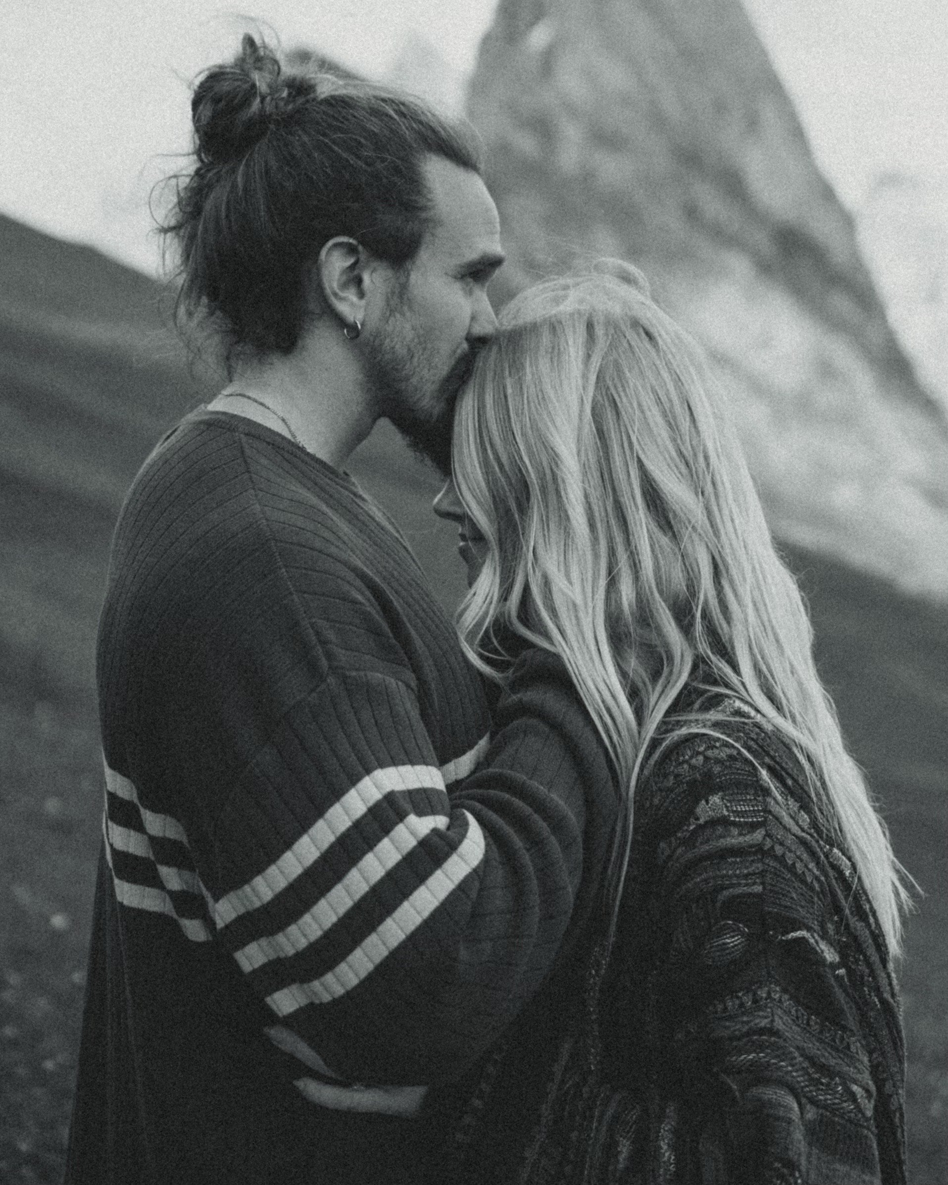 A black and white photograph of a man and woman in an intimate embrace outdoors. The man has a beard, long hair tied up, and is kissing the woman's forehead. The woman has long, wavy hair and is smiling with her eyes closed. They are dressed in casual clothing, with a mountainous landscape in the background.