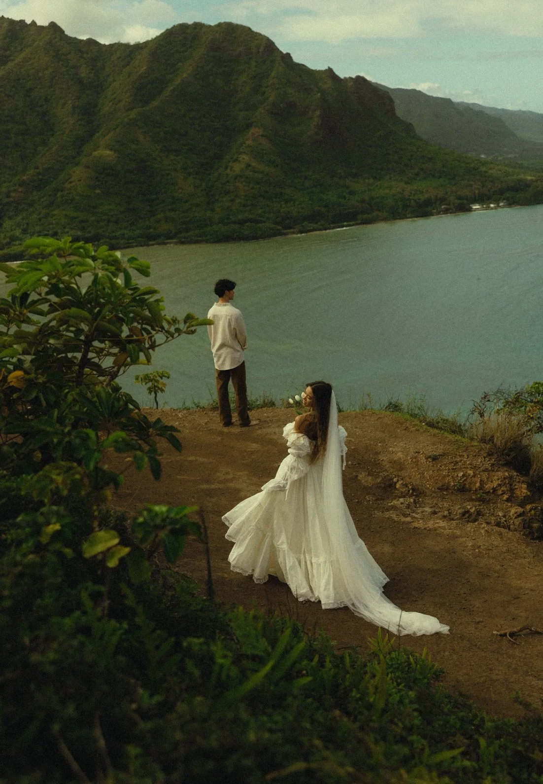 A woman in a white wedding dress holding flowers looks back as a man in a white shirt and brown pants stands near a body of water with green mountains in the background.