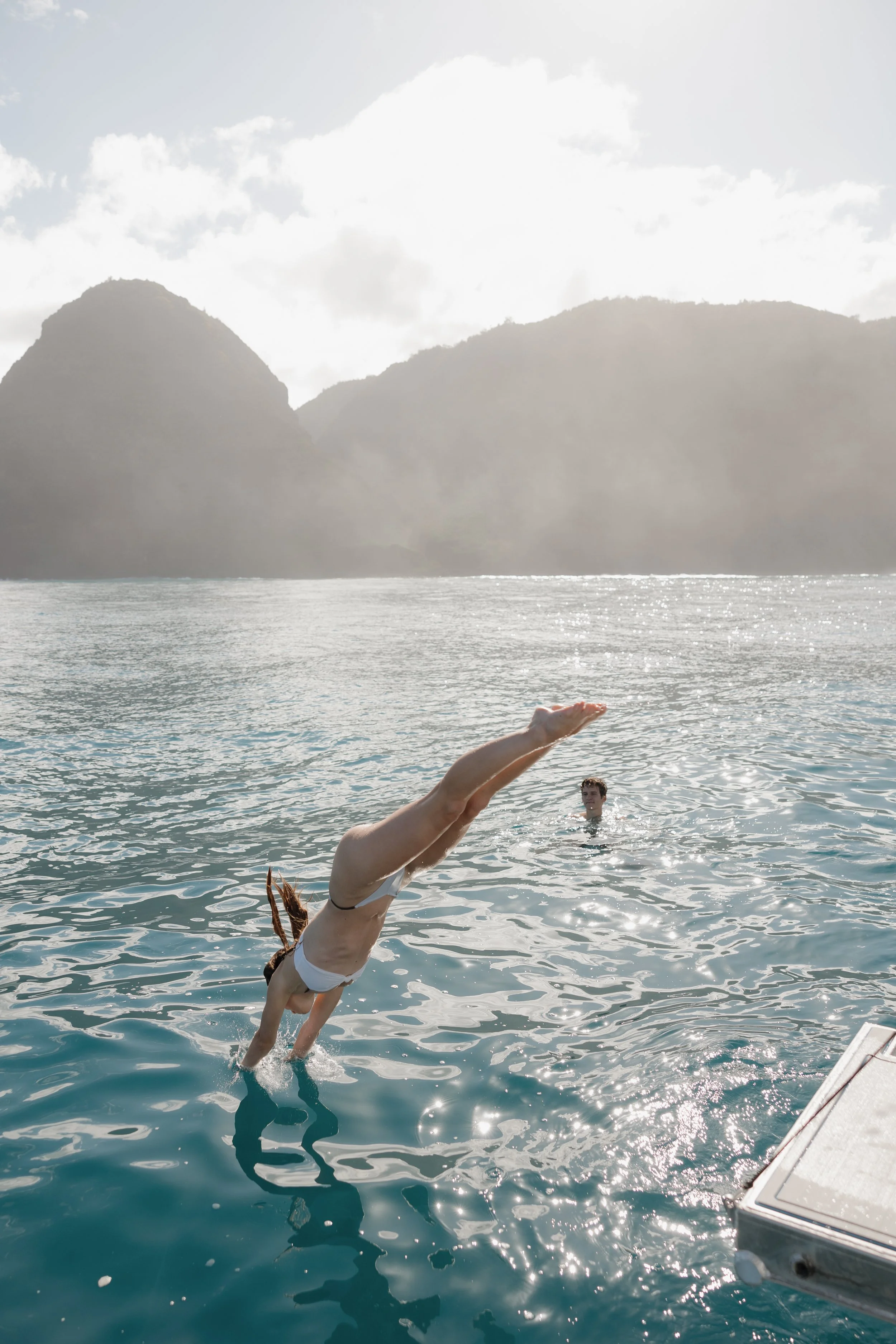 A woman diving into the water from a boat with a man swimming nearby, with mountains and cloudy sky in the background.