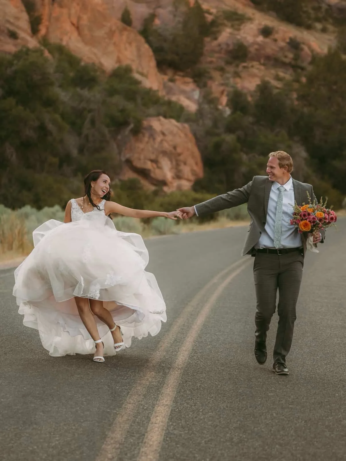A bride and groom holding hands and dancing on a deserted road in a scenic mountain landscape
