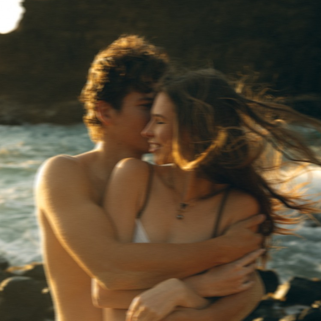 A couple embracing and smiling at each other on a rocky beach with ocean waves in the background. The image is slightly blurred.