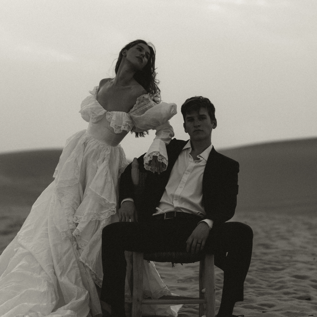 Black and white photo of a woman in a vintage dress standing next to a man in a suit seated on a wooden chair, in a desert landscape.