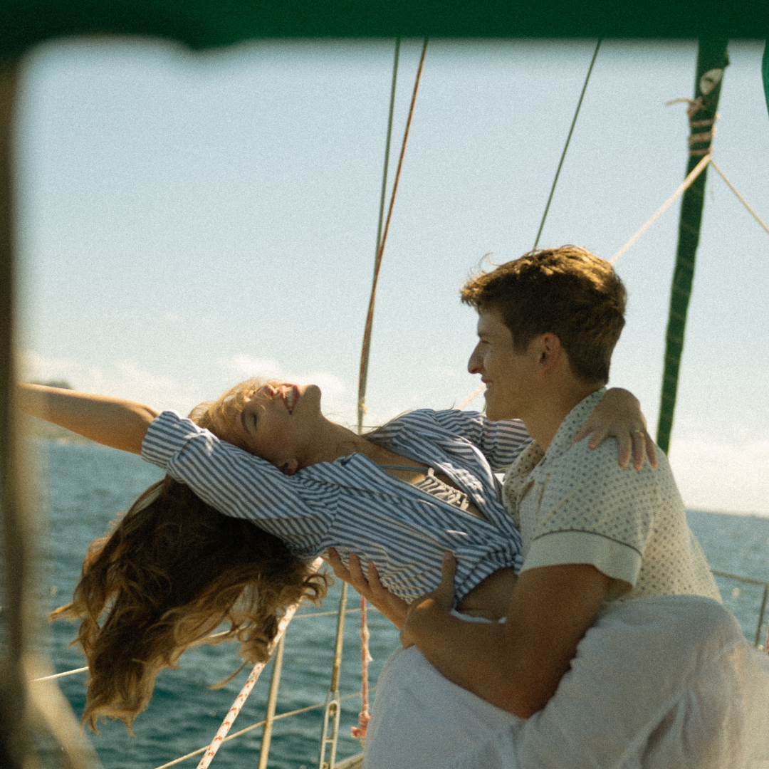 A young man holding a woman on a boat, with the woman stretching her arms out and smiling, against a backdrop of water and a clear sky.