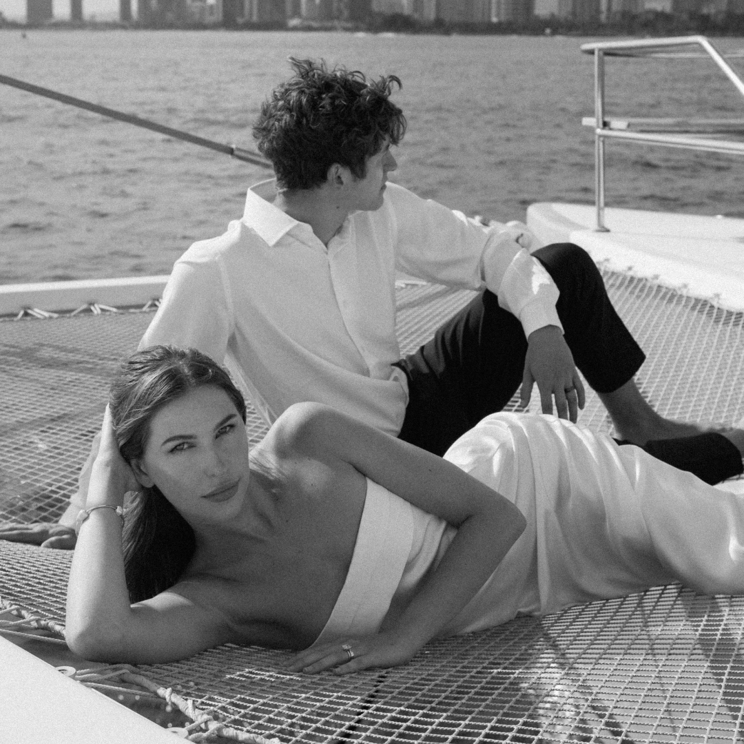 A man and a woman relax on a boat, with the woman lying on her side and the man sitting behind her, both looking away from each other. The scene is outdoors with water and city skyline in the background.