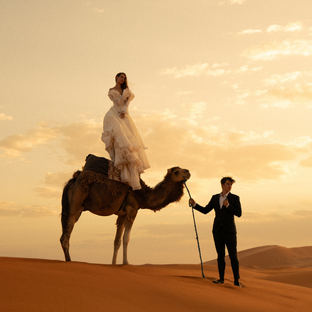 A model in a white wedding dress rides a camel in a desert at sunset, with a man holding the camel's leash.
