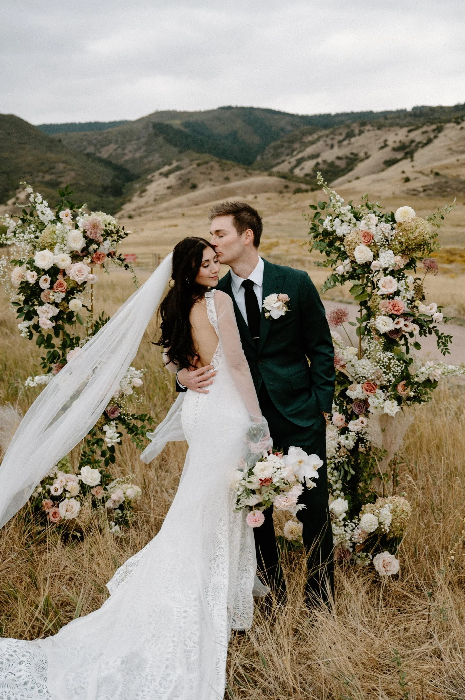 A bride and groom in wedding attire sharing a kiss outdoors next to floral arch and bouquet in a natural, mountain-filled landscape.