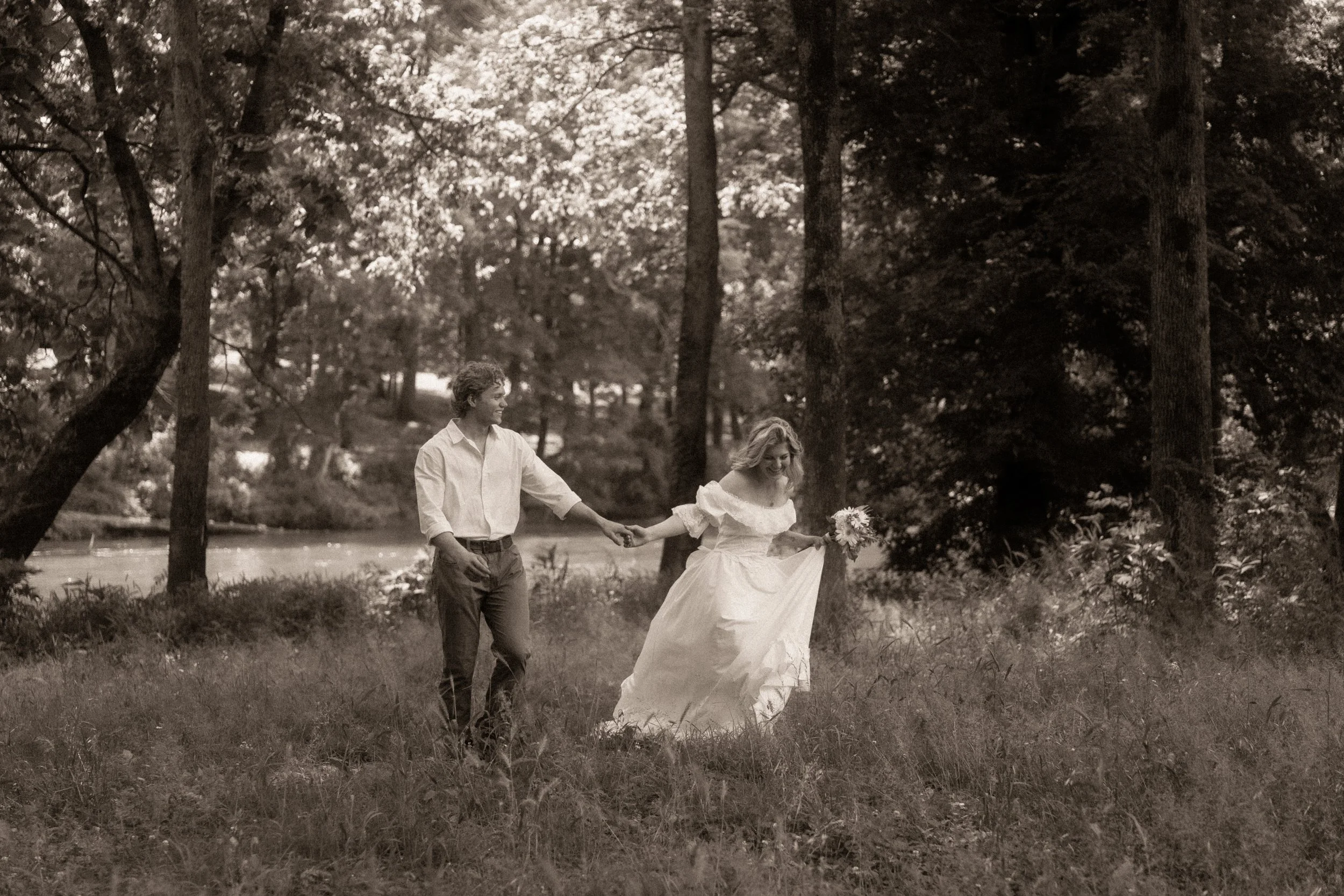 A black and white photo of a bride and groom walking hand in hand through a wooded area, holding a bouquet, with tall trees and grass around them.