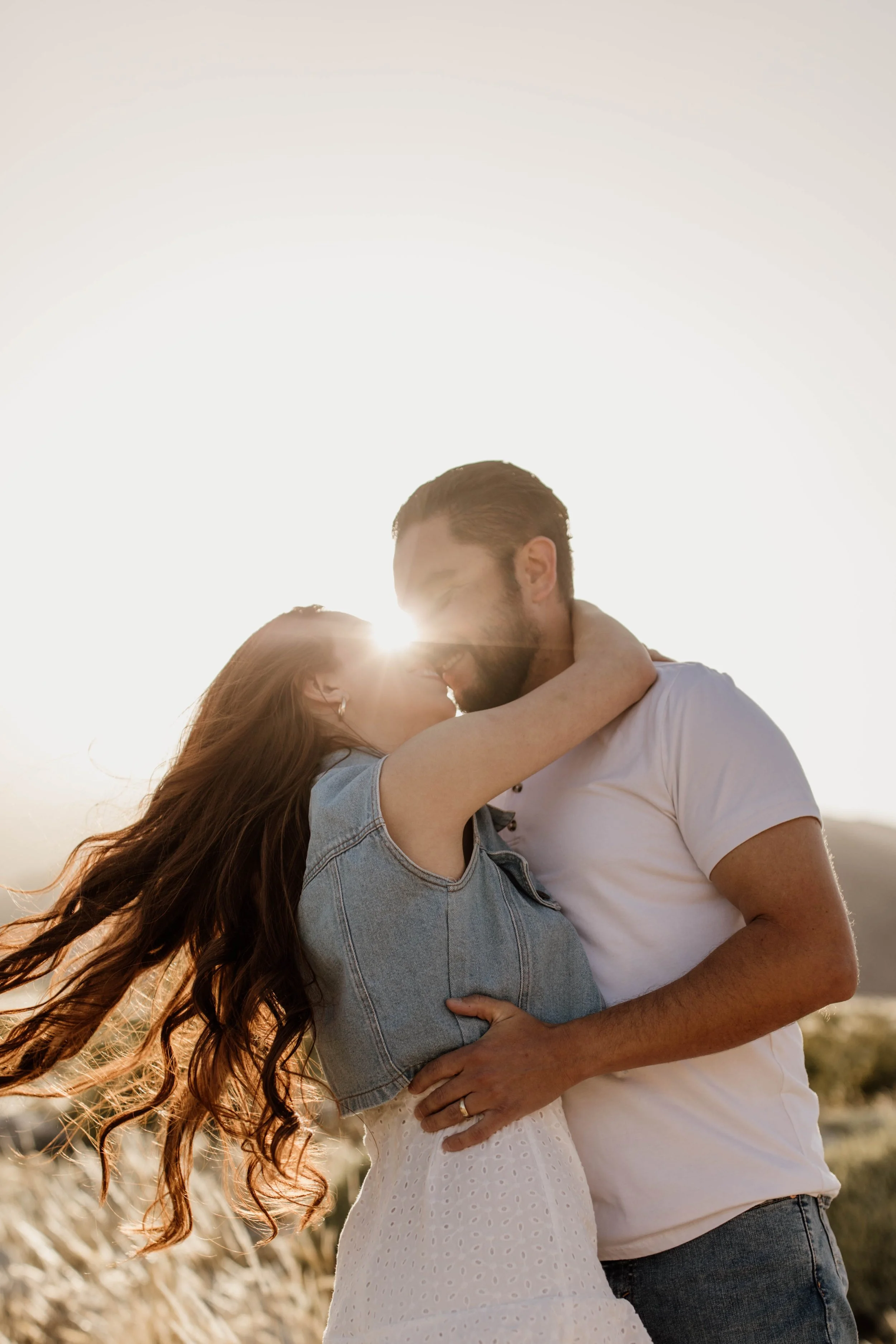 A couple embracing and kissing outdoors during sunset, with the sun shining behind them.