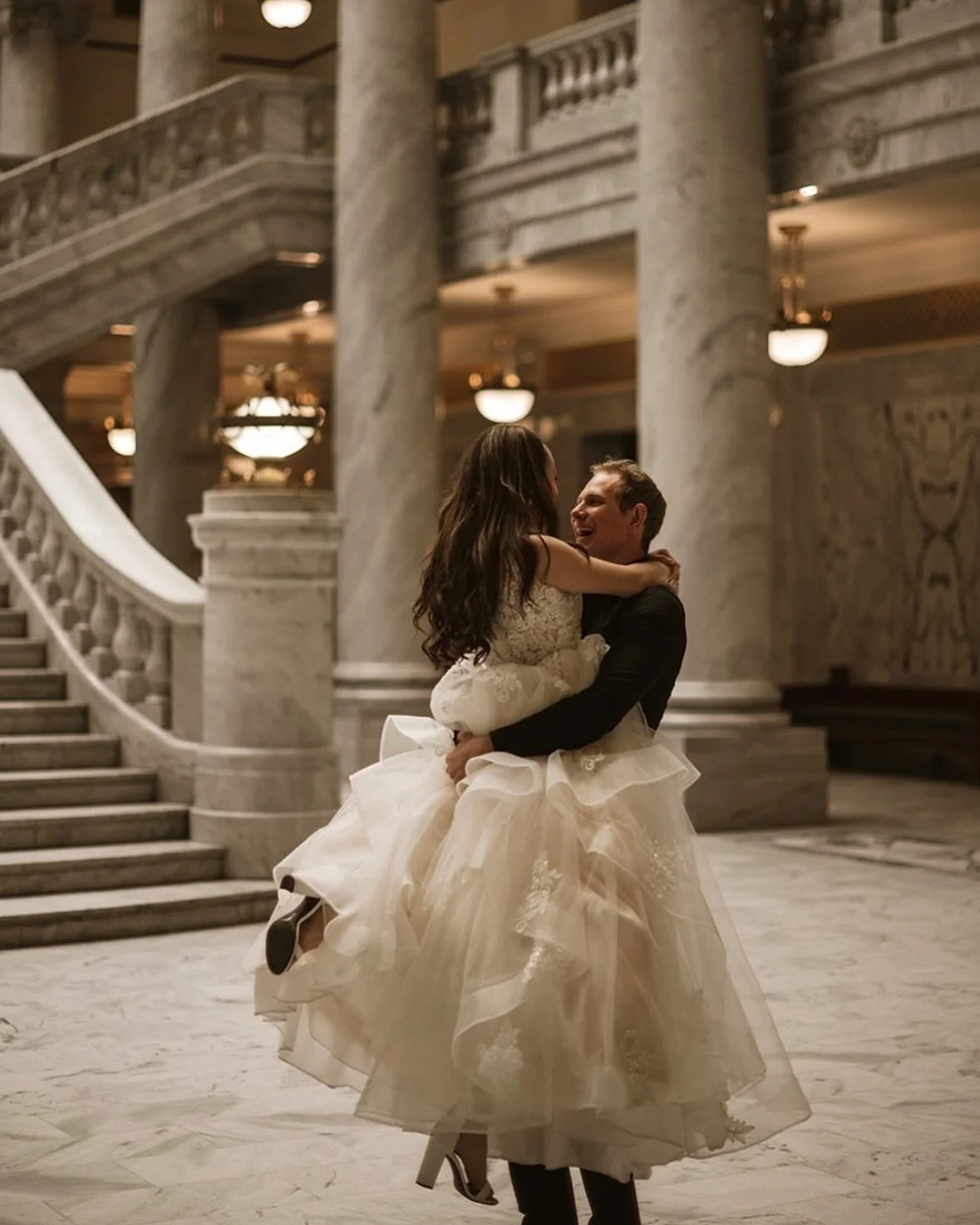 A man in a dark suit lifts a woman in a wedding dress in a grand marble building, both smiling, with marble stairs and columns in the background.