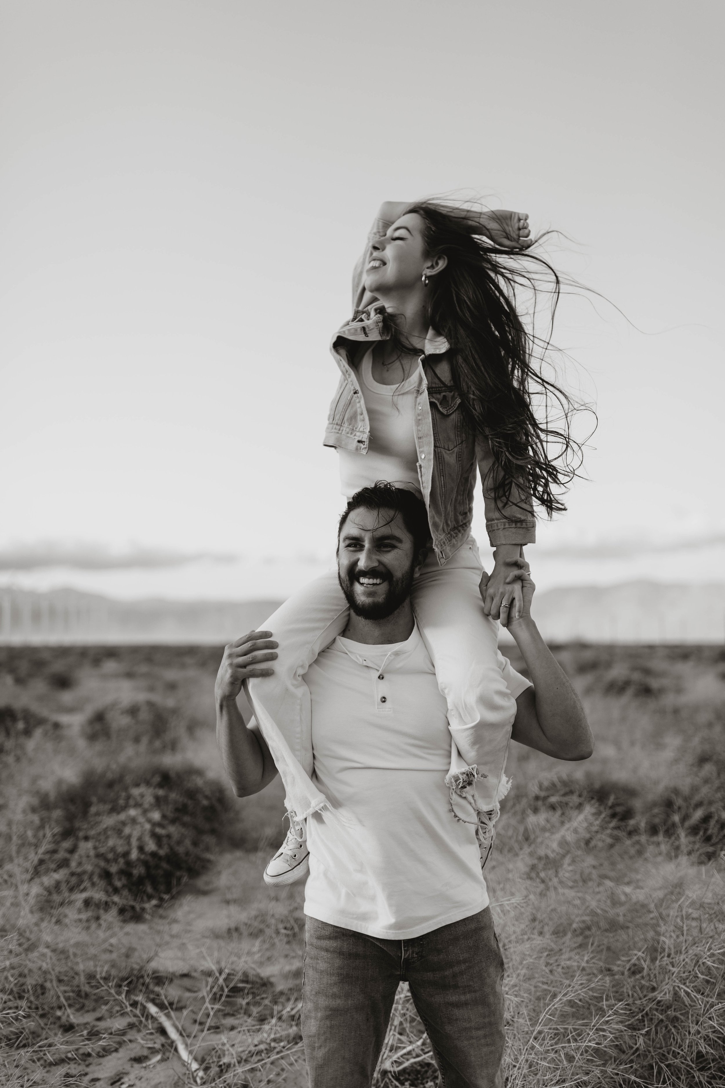 A man giving a woman a piggyback ride on a sandy, grassy path outdoors, with a wide open landscape in the background, black and white photo.