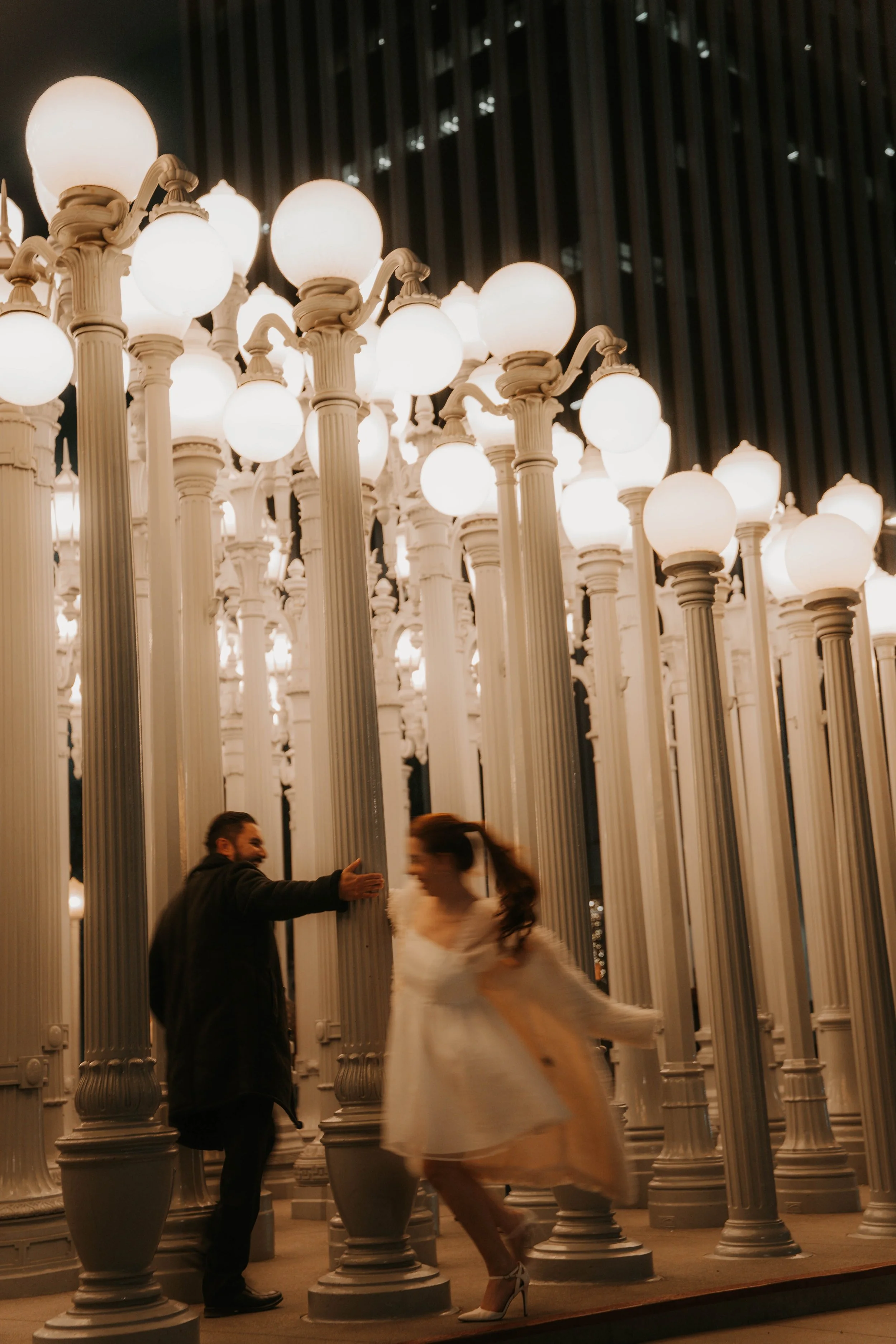 Two people, a man and a woman, are interacting near tall streetlamp-like structures with glowing globes in an urban setting at night.