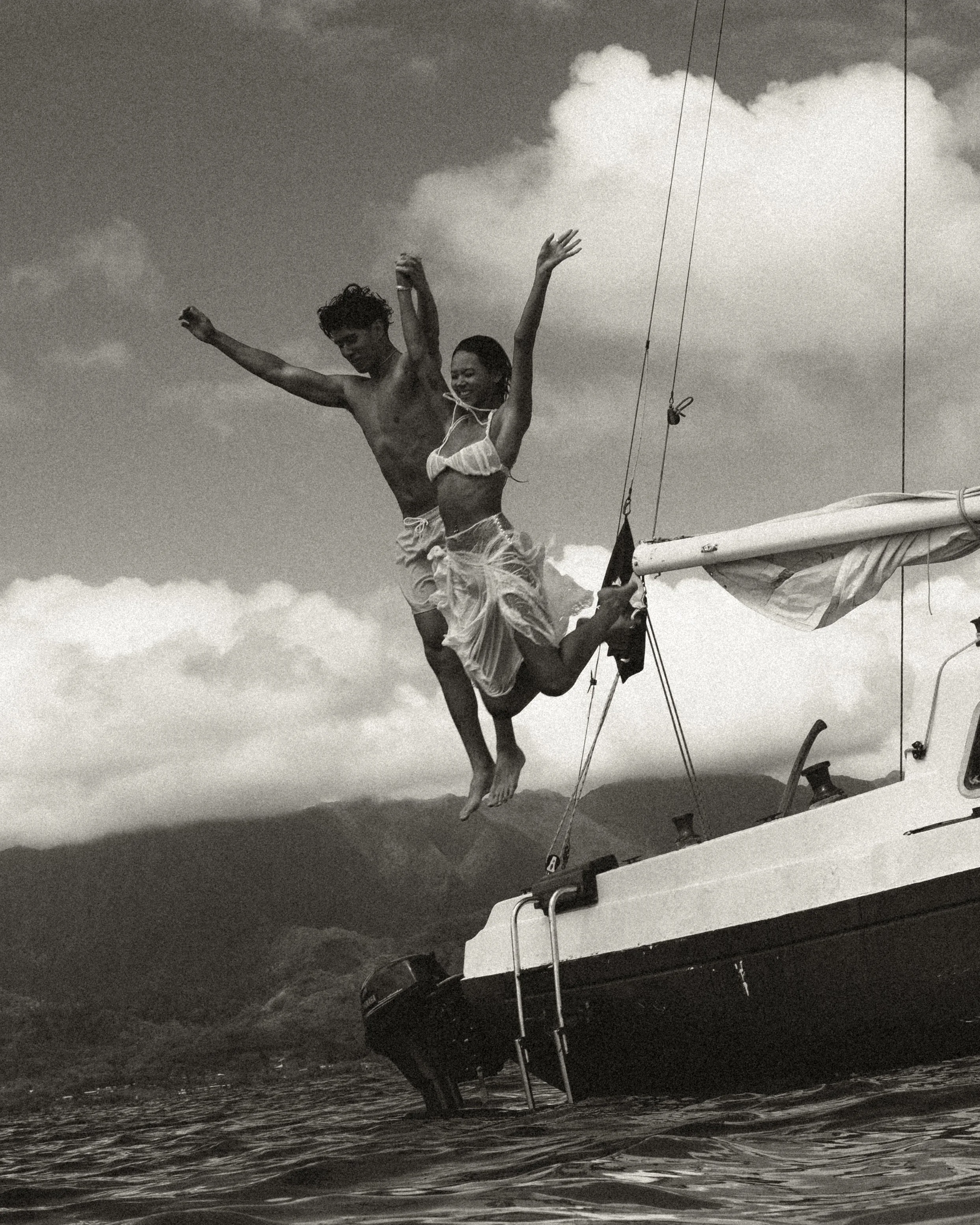A black-and-white photo of a man and woman jumping off a boat into the water with mountains in the background.