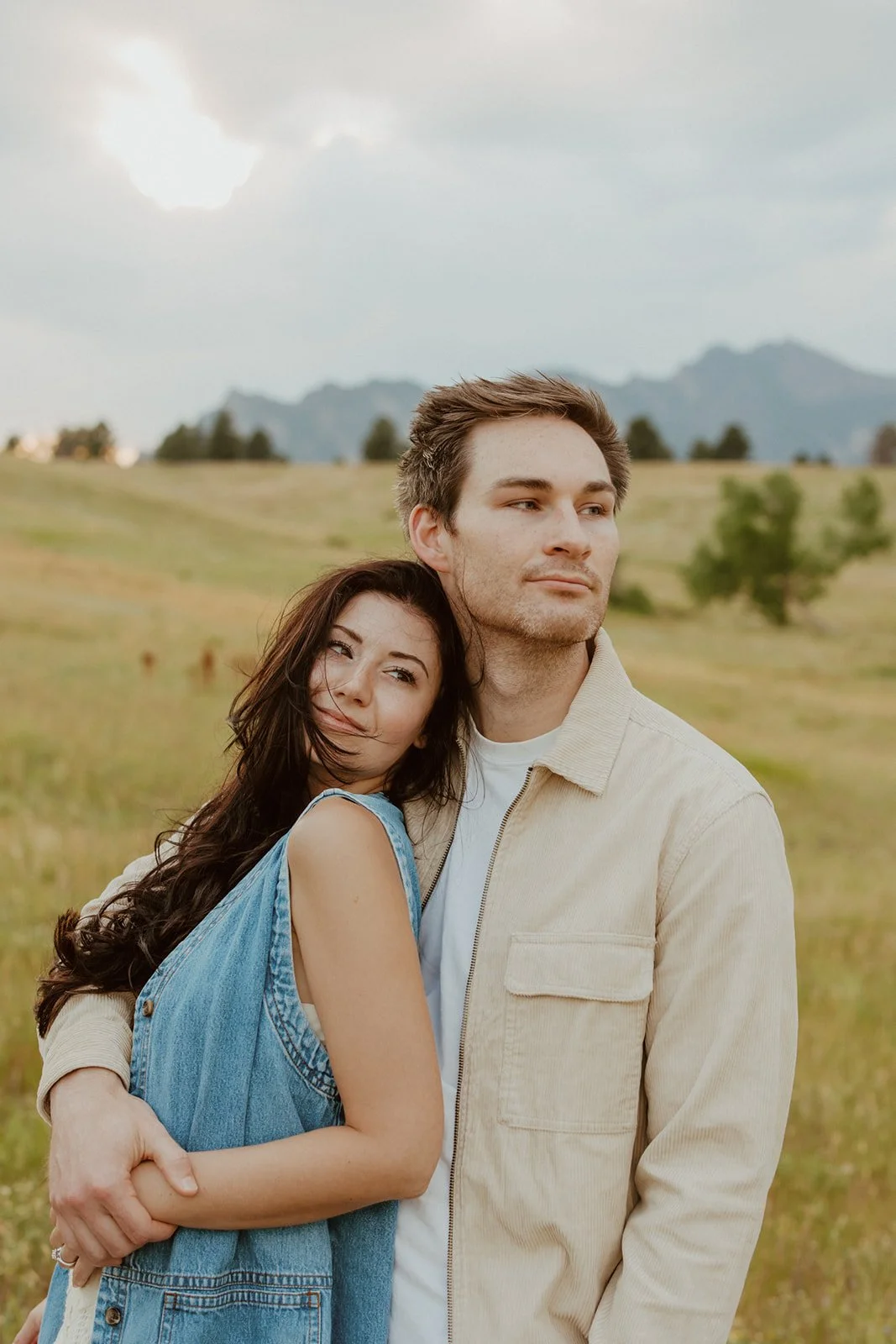 A young woman with long dark hair hugging a young man with short brown hair, both standing outdoors in a grassy field with hills and mountains in the background during daytime.