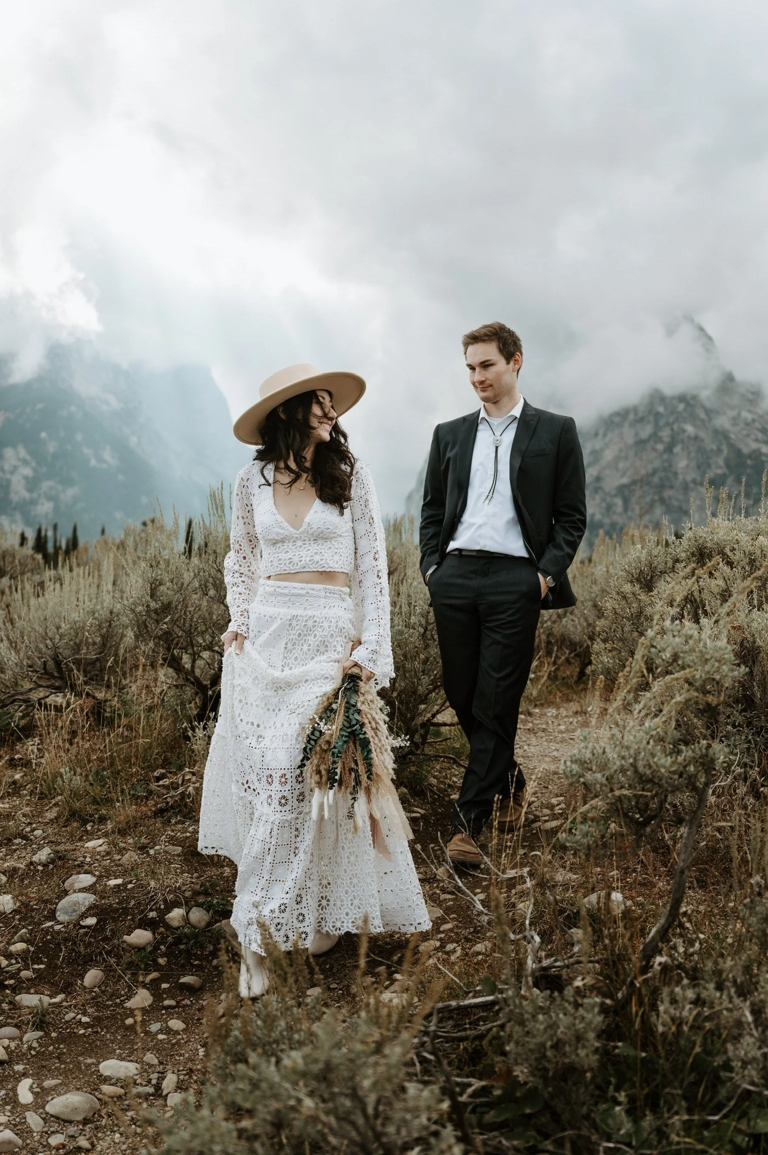 A woman in a white bohemian-style dress and wide-brimmed hat holds a bouquet of flowers, standing on a dirt trail with a man in a black suit nearby in a mountainous landscape with cloudy sky.