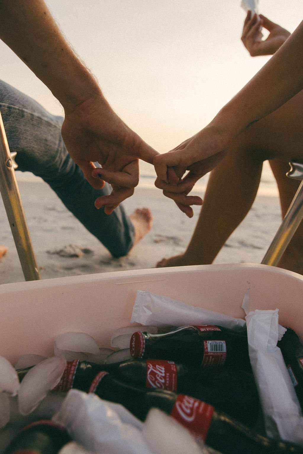 Two people holding hands on a beach with a container of ice and Coke bottles in the foreground.