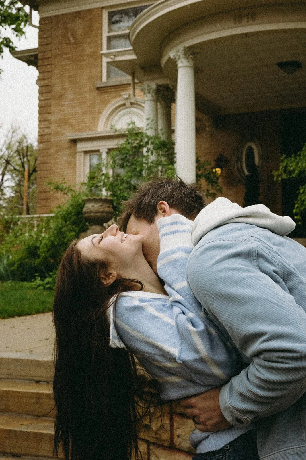 A couple sharing a kiss outside a brick house with white columns and lush greenery.
