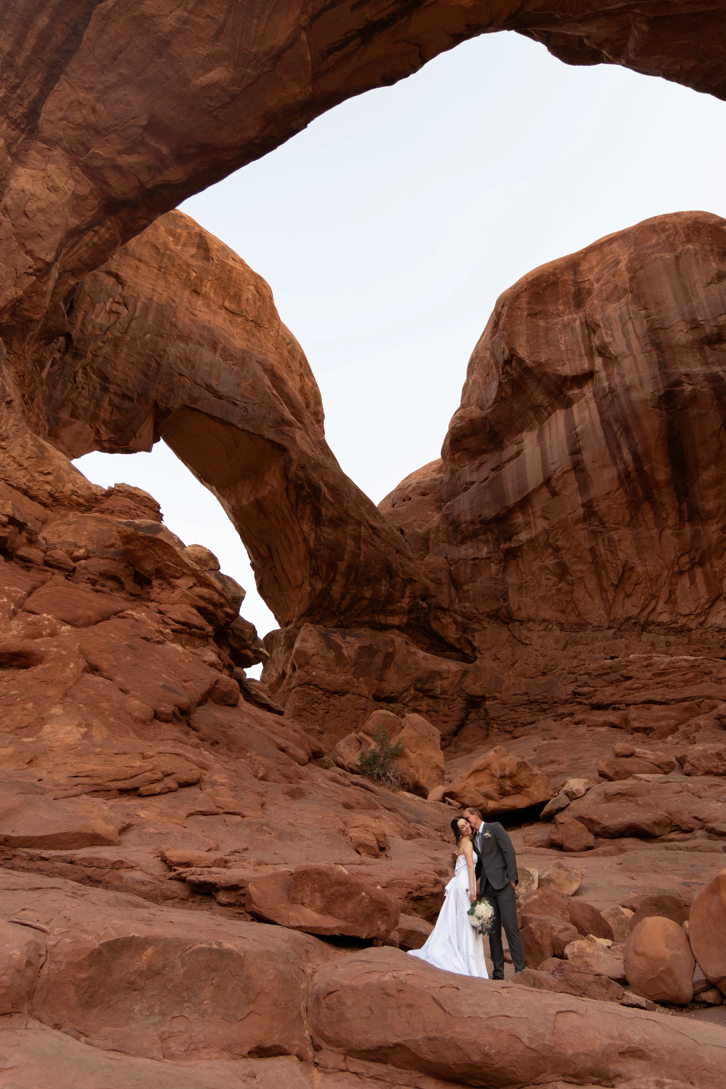 A bride and groom in wedding attire stand close together in a desert canyon with red rock formations and natural arches.