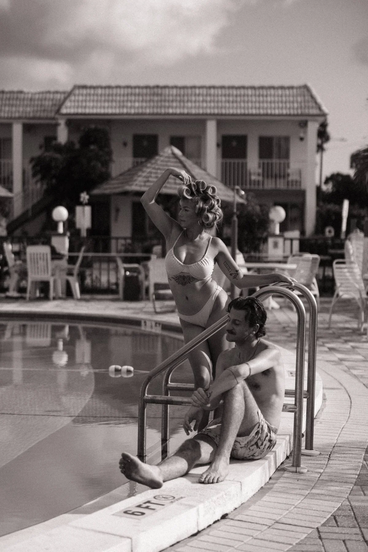 A woman with vintage styled hair curling rollers and a swimsuit stands on the poolside ladder, while a man sits nearby with a towel around his waist, at a hotel pool area.