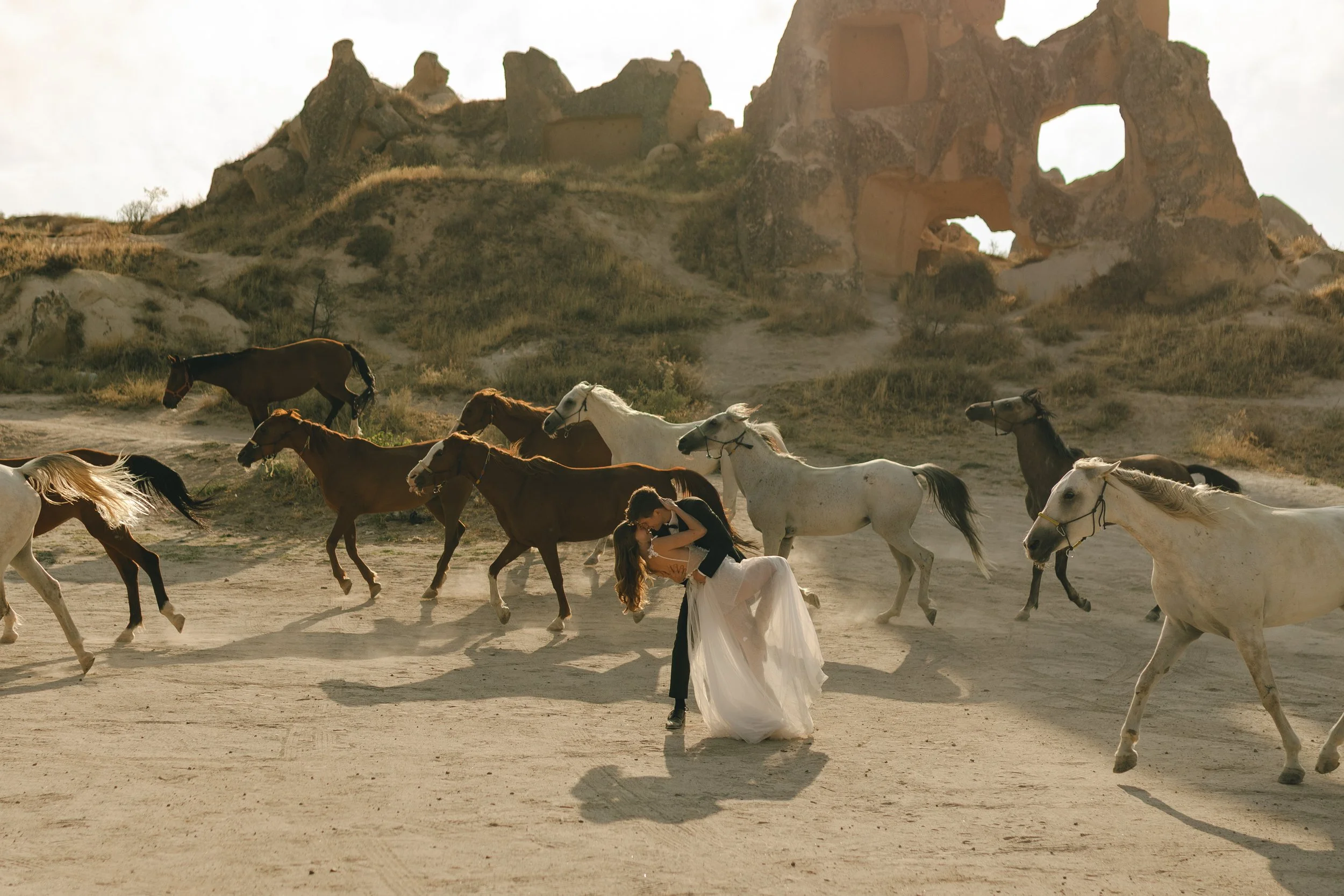 A model couple in formal attire, with the woman in a white wedding dress and the man in a suit, embracing and kissing while standing on a dusty path surrounded by running horses, with rocky hills and cave dwellings in the background.