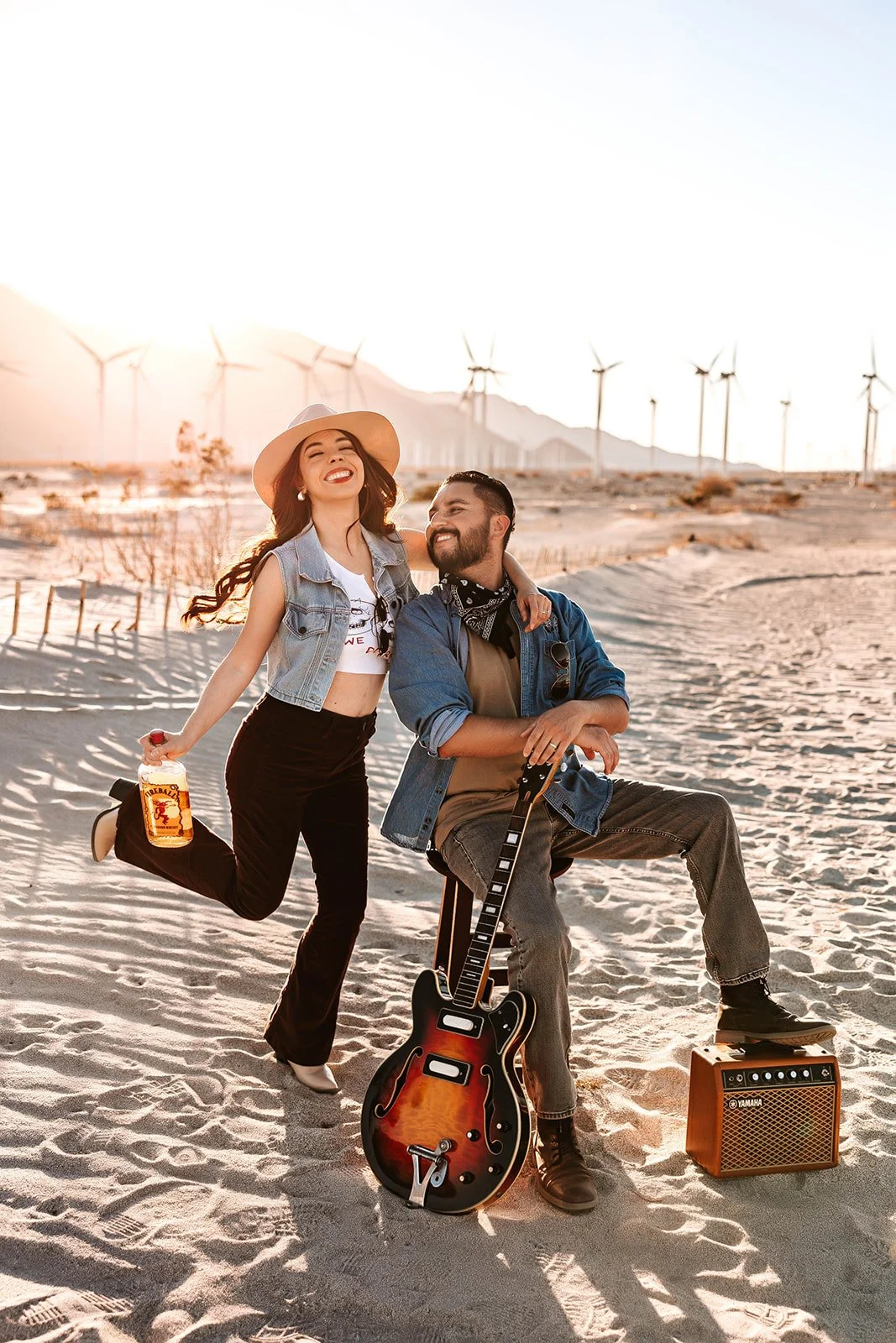 A smiling woman with long hair wearing a hat and jeans vest standing on one leg, holding a bottle. A man with a beard, sitting with one foot on a small amplifier and the other on the ground, holding an electric guitar on a sandy landscape with wind turbines in the background, during sunset.