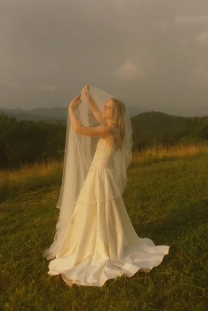 A woman in a white dress standing outdoors on a grassy field, holding a sheer veil above her head, with a landscape of hills and dark clouds in the background.