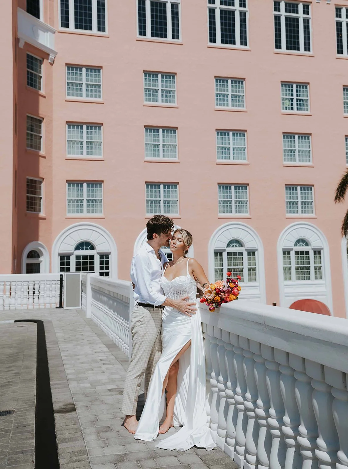 A newlywed couple standing on a white bridge in front of a pink building with multiple windows, the bride holding a colorful bouquet of flowers.