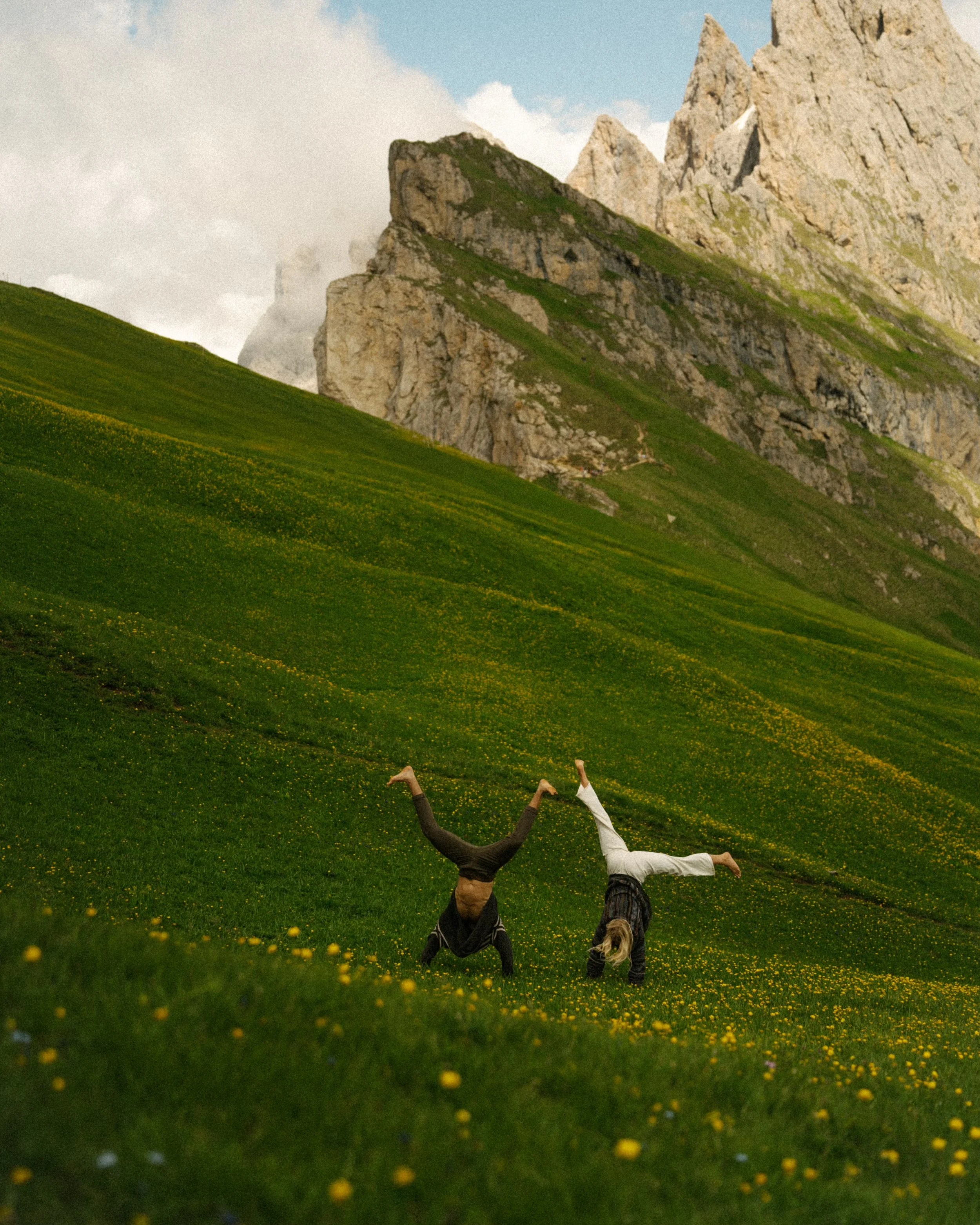 Two individuals performing handstands on a lush green field with yellow flowers in front of mountain peaks under a partly cloudy sky.