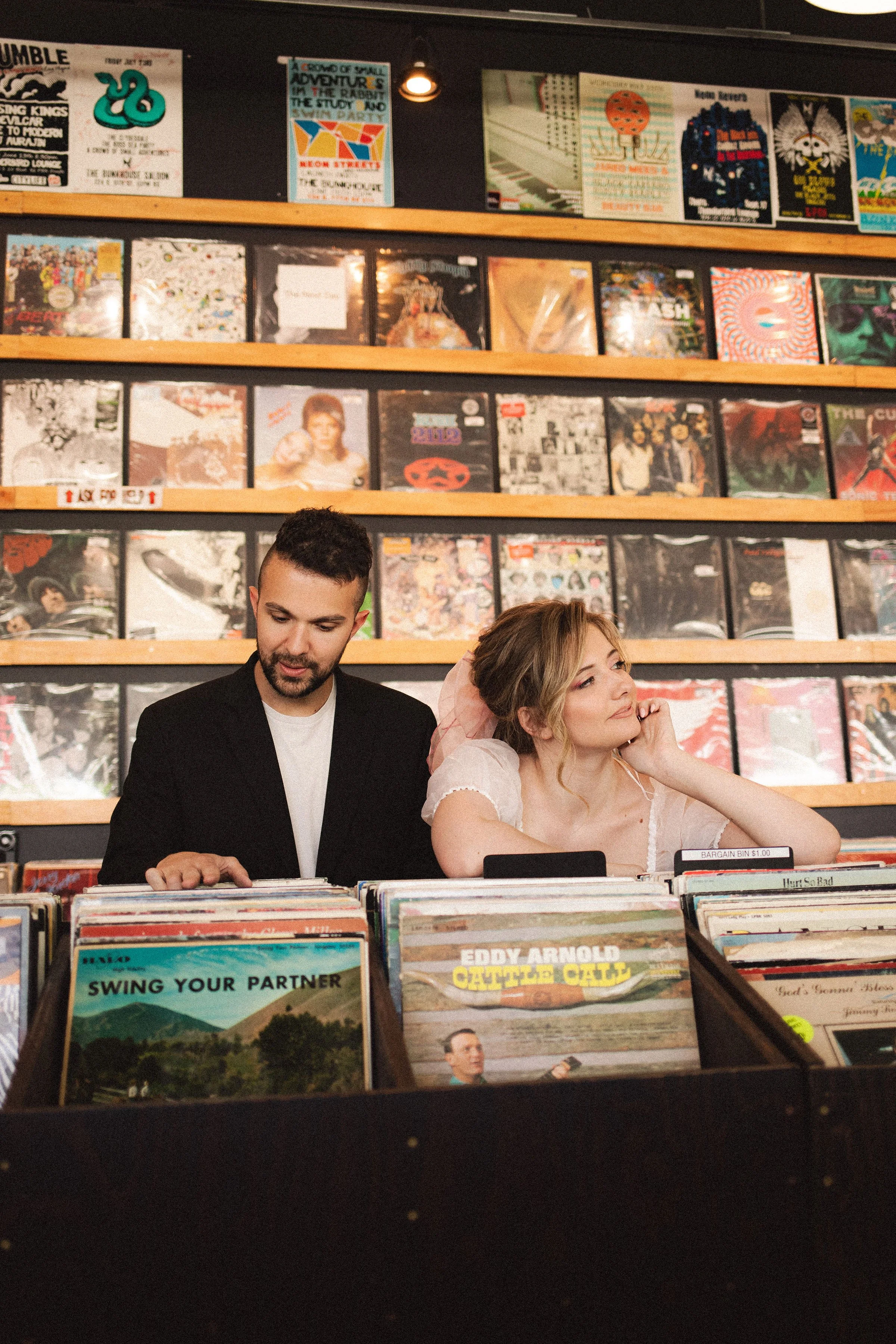 A man and a woman browsing vinyl records in a record store. The woman is dressed as a bride with a pink veil, and the man has a beard and is wearing a black jacket. They are looking at records in a bin with album covers visible in the foreground.