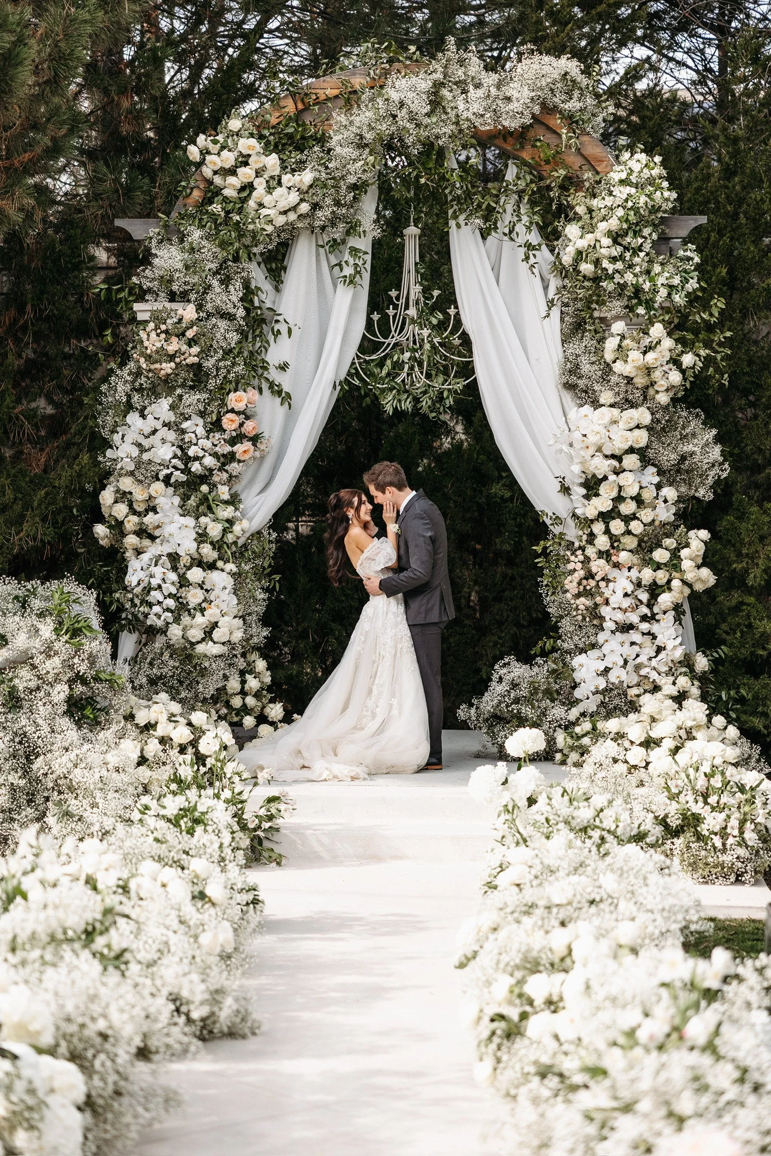 A bride and groom embrace under a wedding arch decorated with white flowers and greenery, with white drapes and a chandelier, on an outdoor wedding stage surrounded by white floral arrangements.