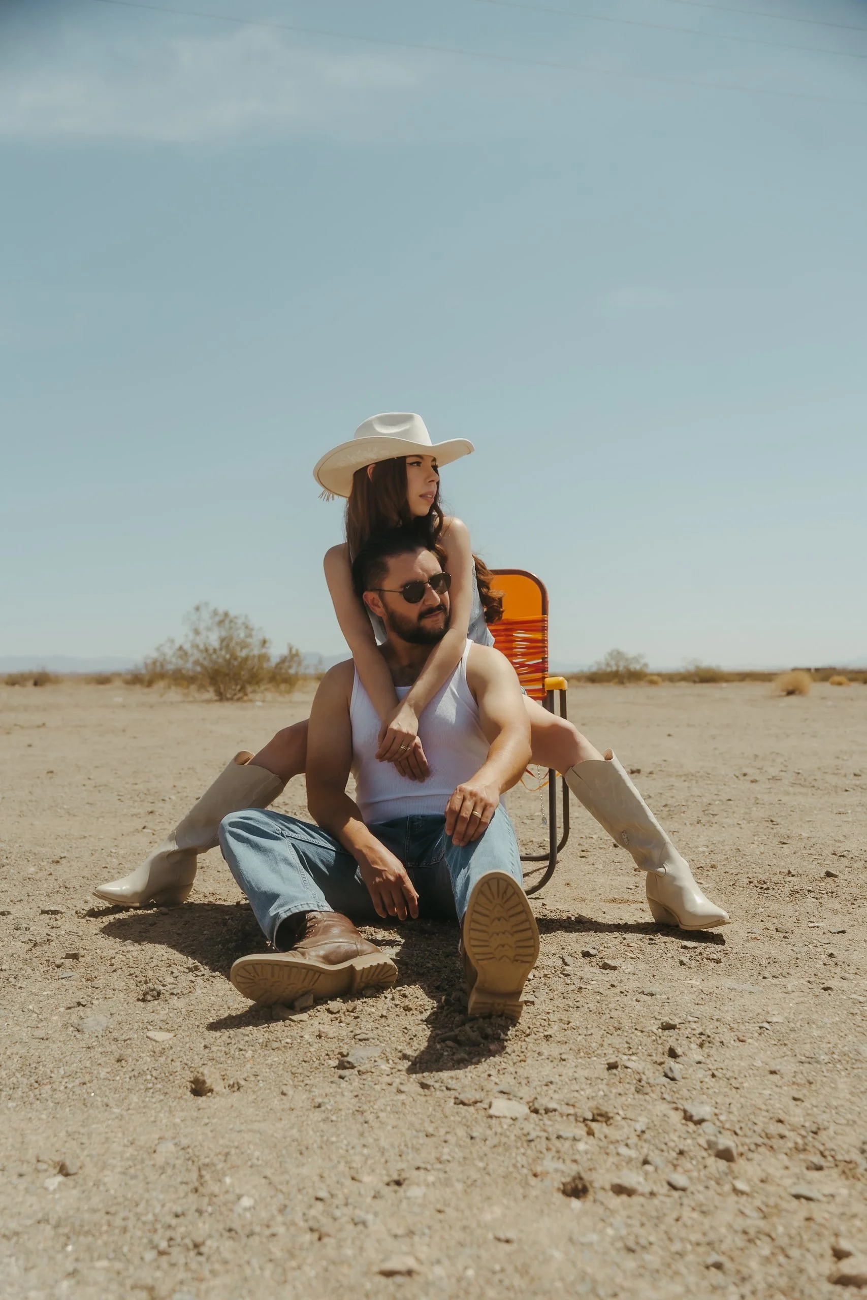 A man and woman sit close together in a desert landscape. The woman wears a wide-brimmed hat and knee-high boots, sitting behind the man who has sunglasses and a tank top. They appear relaxed and close to each other.