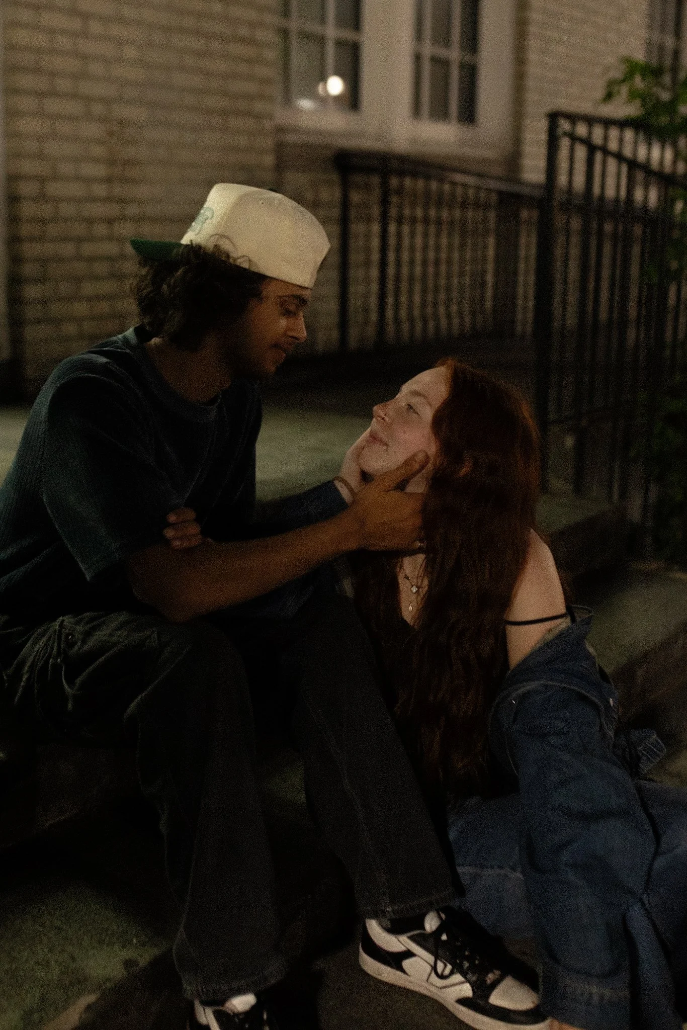 A young man with long hair wearing a white cap looks at a young woman with long red hair, sitting on the ground, touching her face gently, at night outside a building.