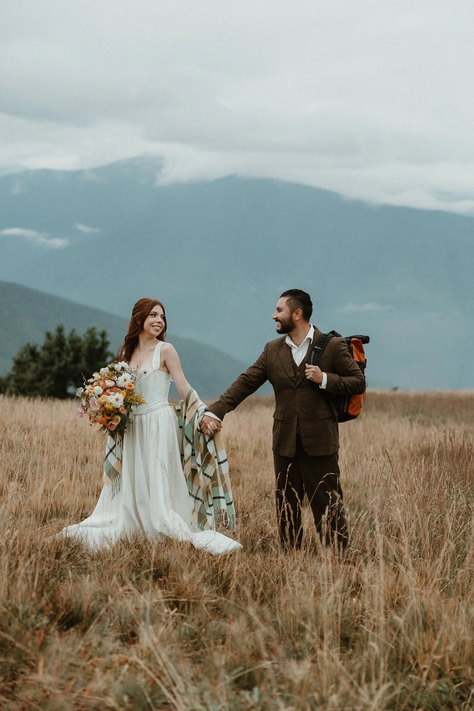 A couple in wedding attire holding hands in a grassy field with mountains in the background.