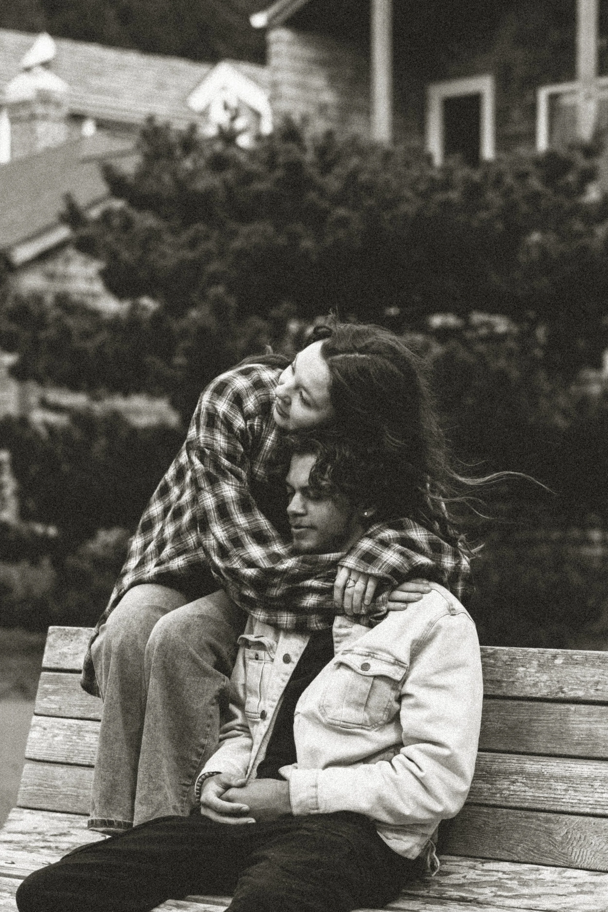 Two young people sitting on a wooden bench outdoors, embracing each other with one on their lap, in front of a large tree and houses in the background, black and white photograph.
