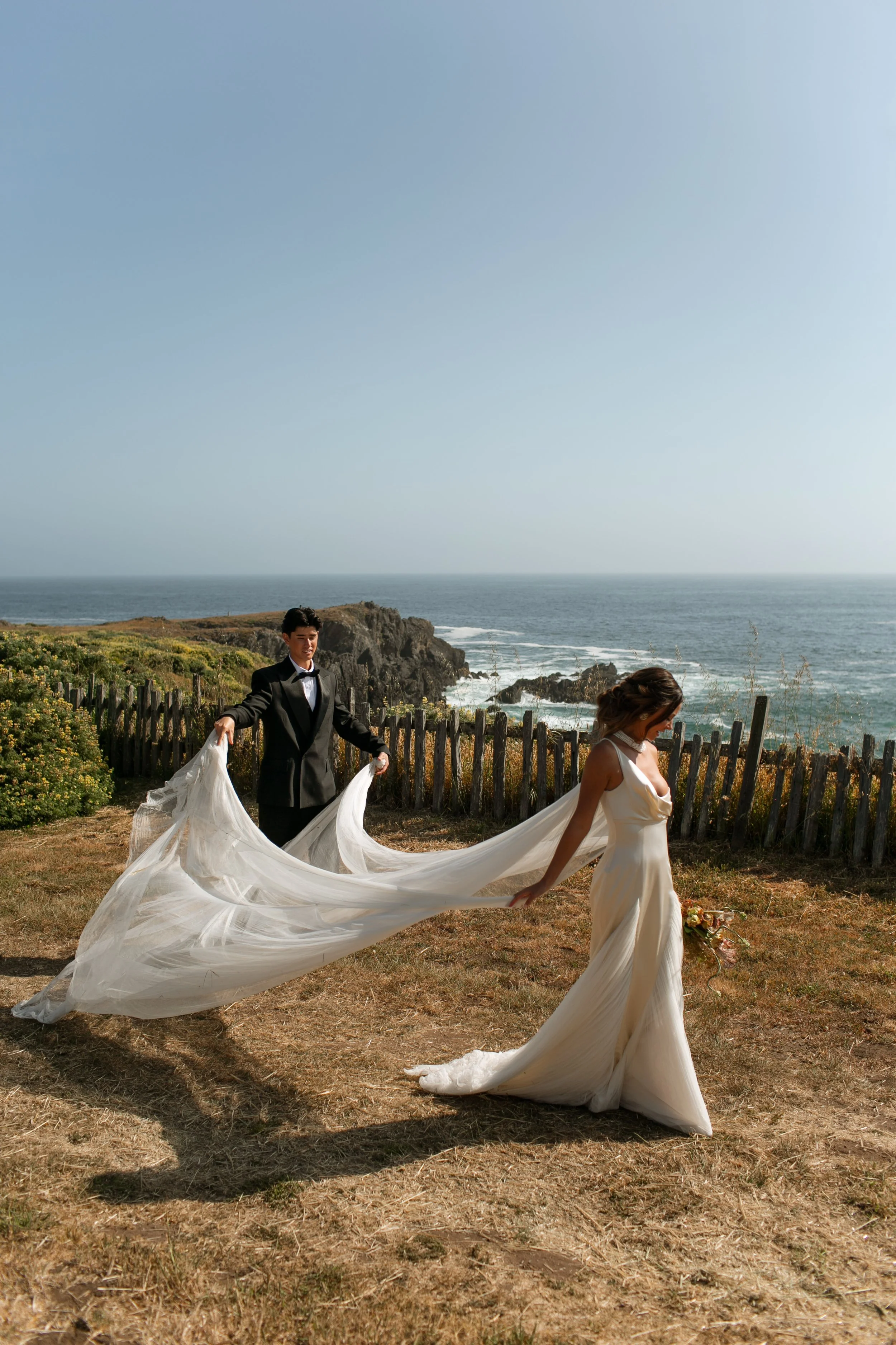 A bride and groom holding a flowing white wedding dress on a grassy area by the ocean, with rocky cliffs and a wooden fence in the background.