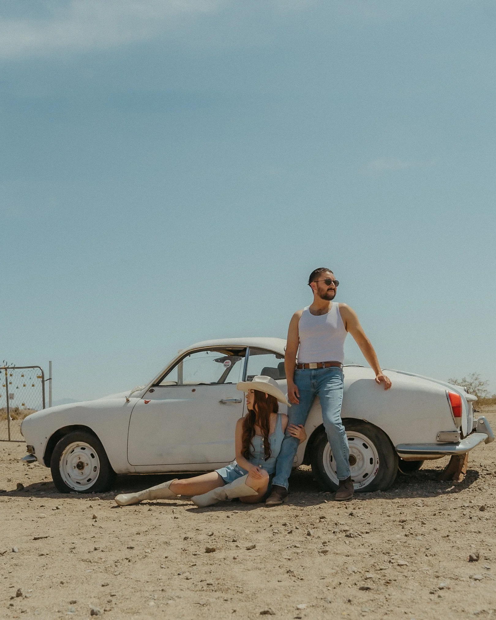 A man and woman with vintage clothing and accessories posing next to a classic white car in a desert landscape under a blue sky.