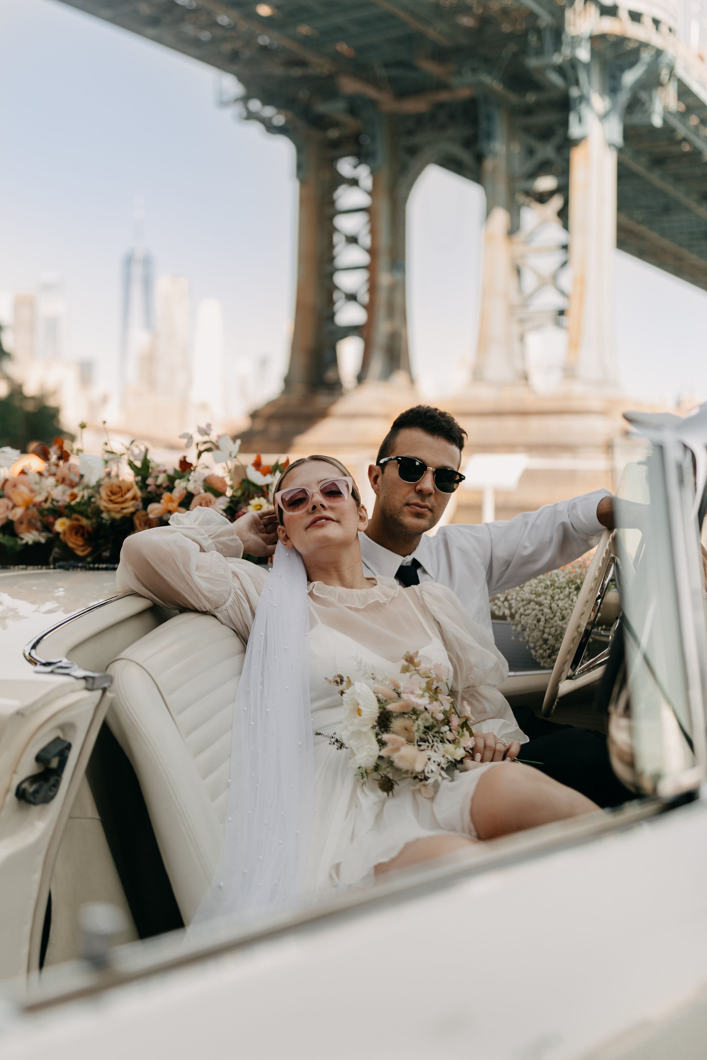 A couple in vintage car, woman in white dress with veil and sunglasses, man in white shirt with black tie and sunglasses, under a bridge with city skyline in background.
