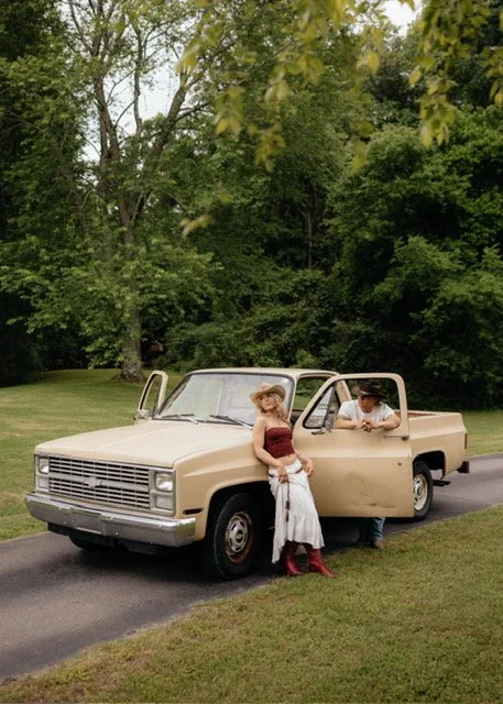 A woman in a strapless red top and white skirt standing next to an old beige pickup truck parked on a rural road, with a man in a hat sitting inside the truck, surrounded by green trees and grass.
