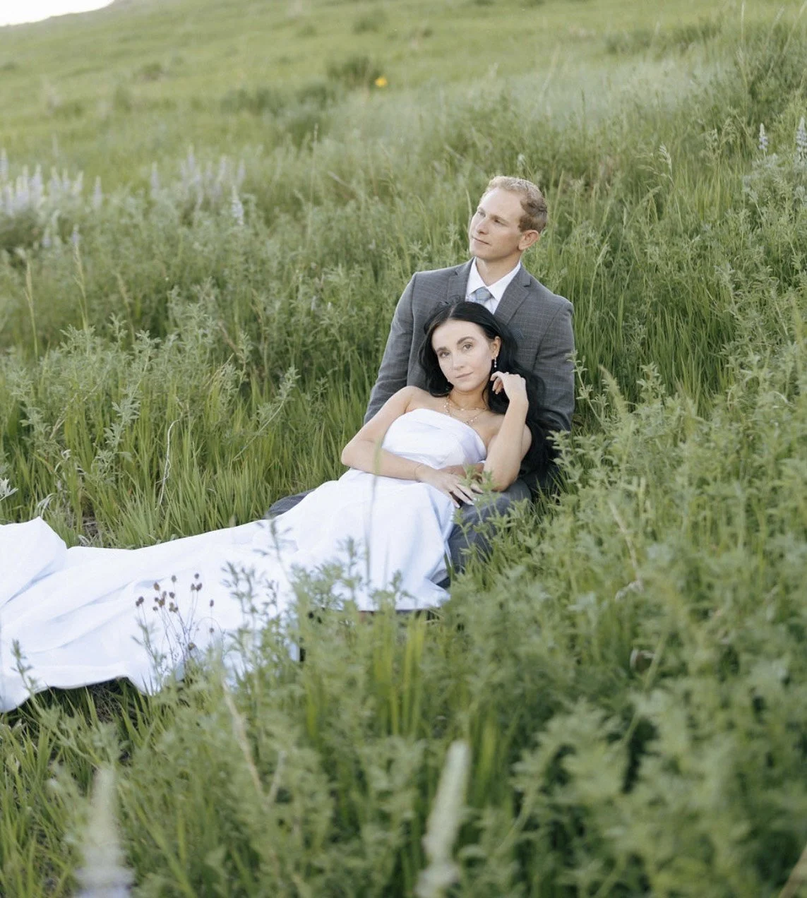 A man and woman sitting together in a field of tall grass with wildflowers. The woman is lying down with her head resting on the man's lap, wearing a white dress, and the man is sitting upright wearing a gray suit. The scene has a peaceful and romantic atmosphere.