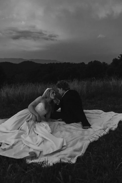 A black and white photo of a couple in wedding attire sitting on a blanket in an open field, facing each other closely with a landscape of trees and hills in the background.