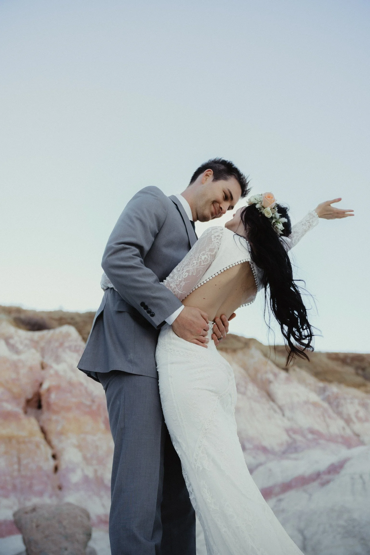 A couple in wedding attire dancing outdoors, with a rocky landscape in the background.