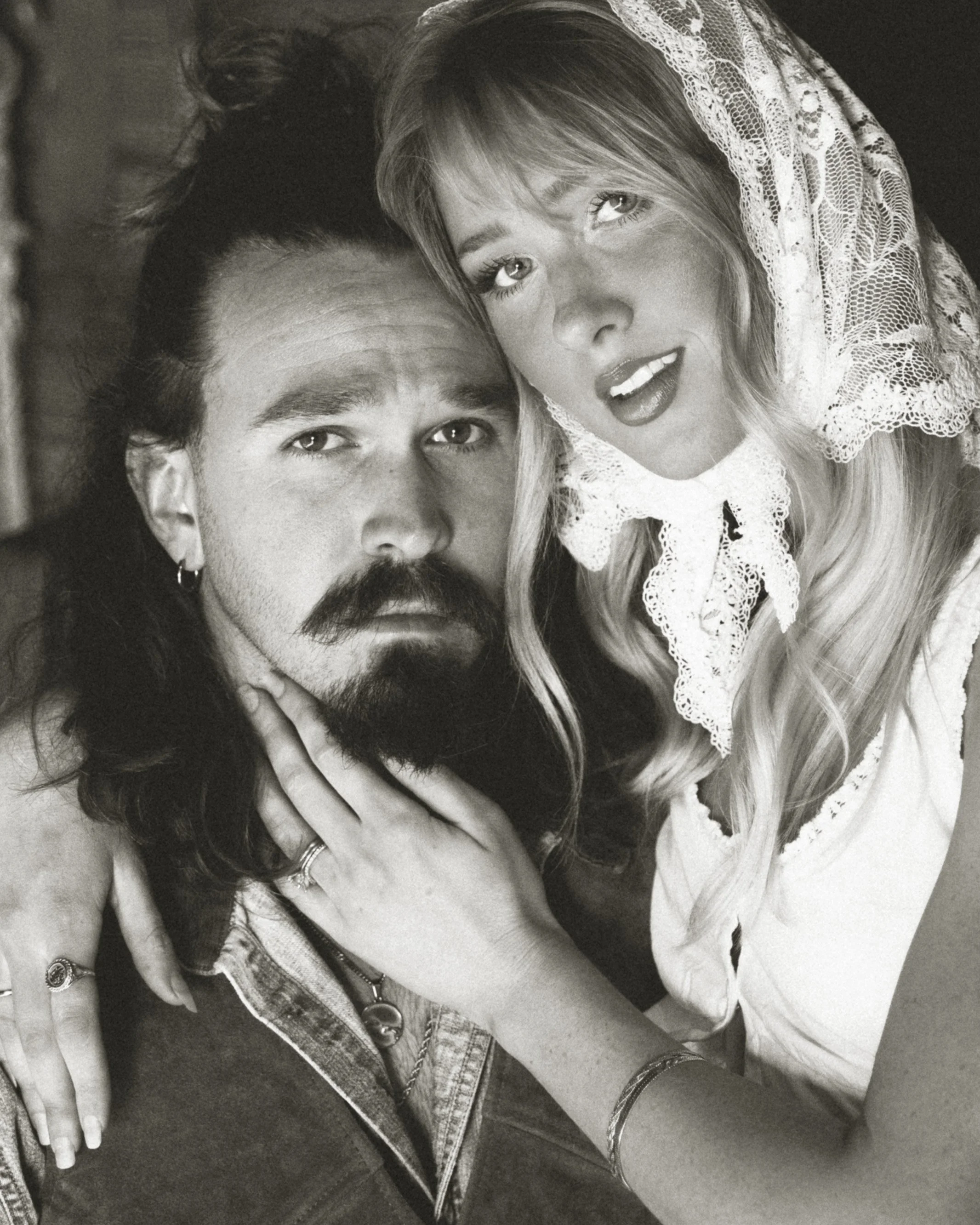 Black and white photo of a man with long dark hair, beard, and mustache, and a woman with long blonde hair wearing a lace bonnet. The woman is leaning close to the man, with her face near his and her hand on his shoulder, both looking at the camera.