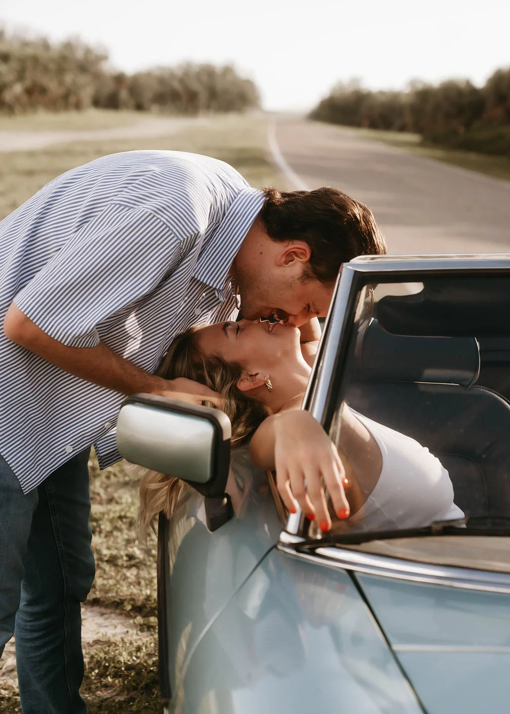 A man and woman sharing an intimate moment next to a car on a rural road in the countryside.