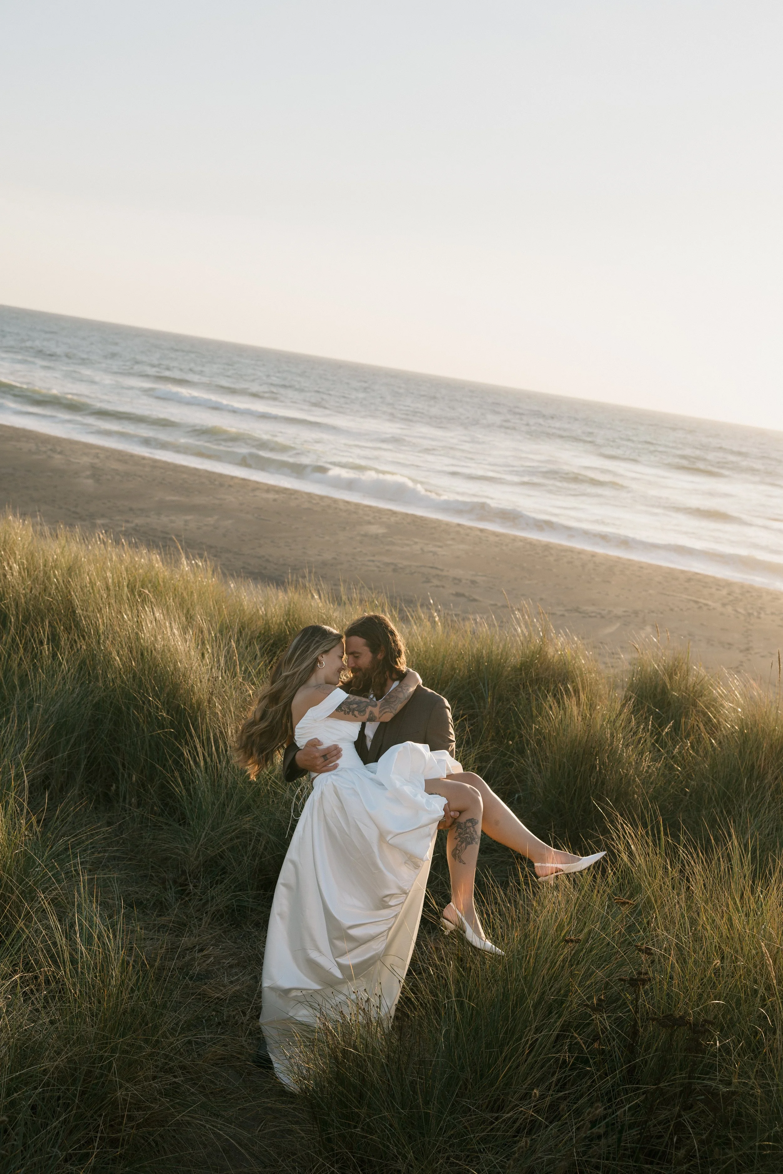 A couple in wedding attire on a grassy dune near the beach, with the ocean and sky in the background, during sunset.
