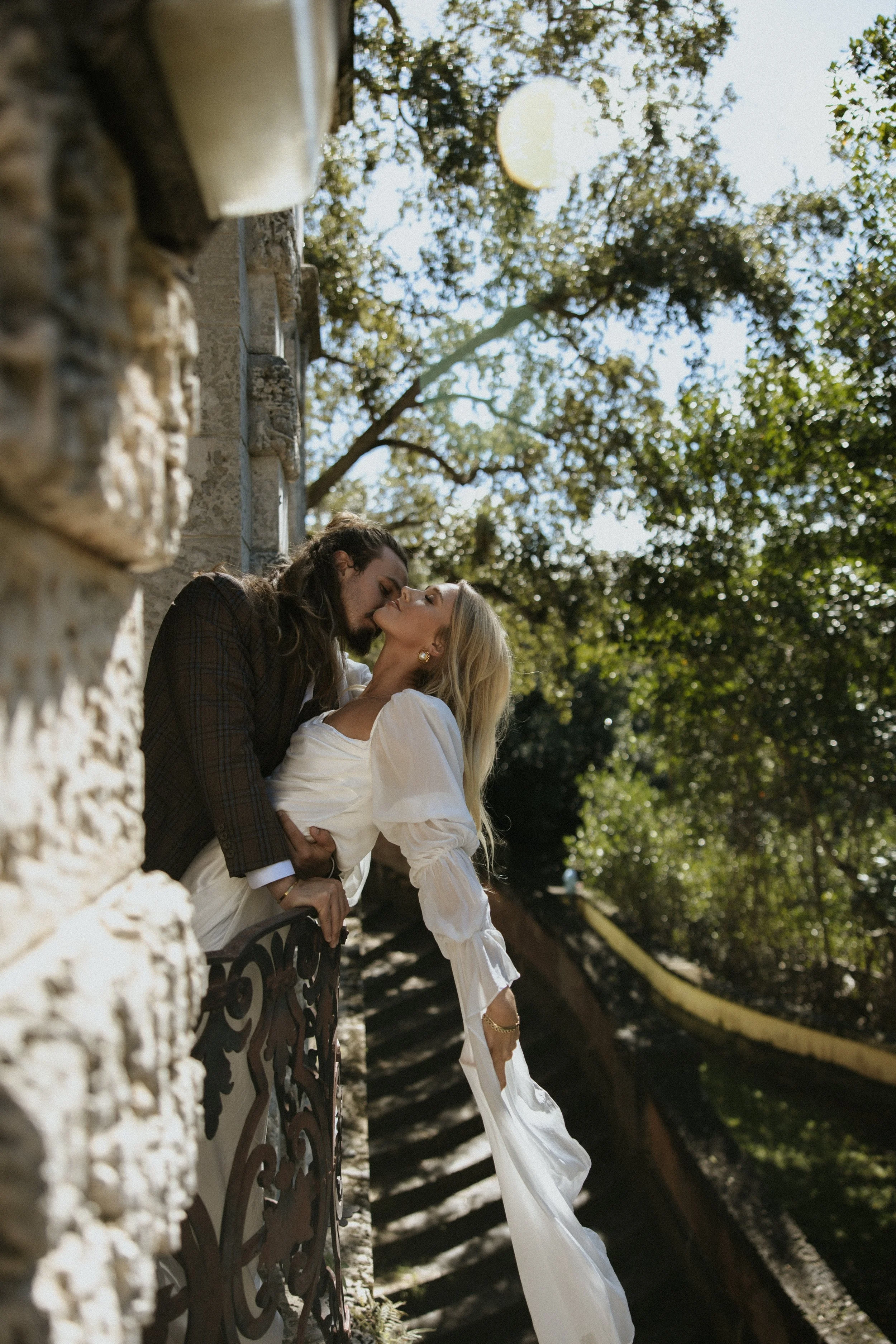 A couple kissing on a balcony with stone walls, trees, and stairs in the background, under a bright sky.